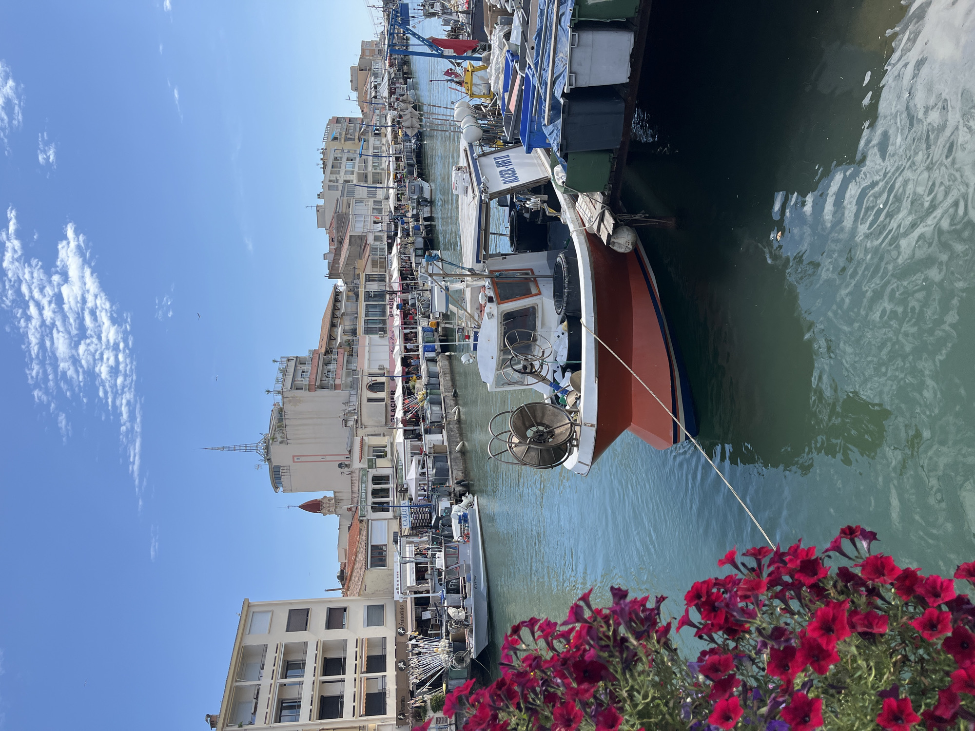 This is a medium shot, taken from a low angle, of a canal in Palavas-les-Flots, France. The canal is lined with buildings and boats. In the foreground, a red and white fishing boat is moored to a dock. It has a winch and fishing gear on deck. Behind it, other fishing boats and a grey boat are also visible. The canal is a vibrant blue-green color, and the sky is clear blue with a few white clouds. The buildings on the canal are a mix of modern and traditional styles, with shops and restaurants on the ground floor. The overall impression is one of a lively and picturesque seaside town.