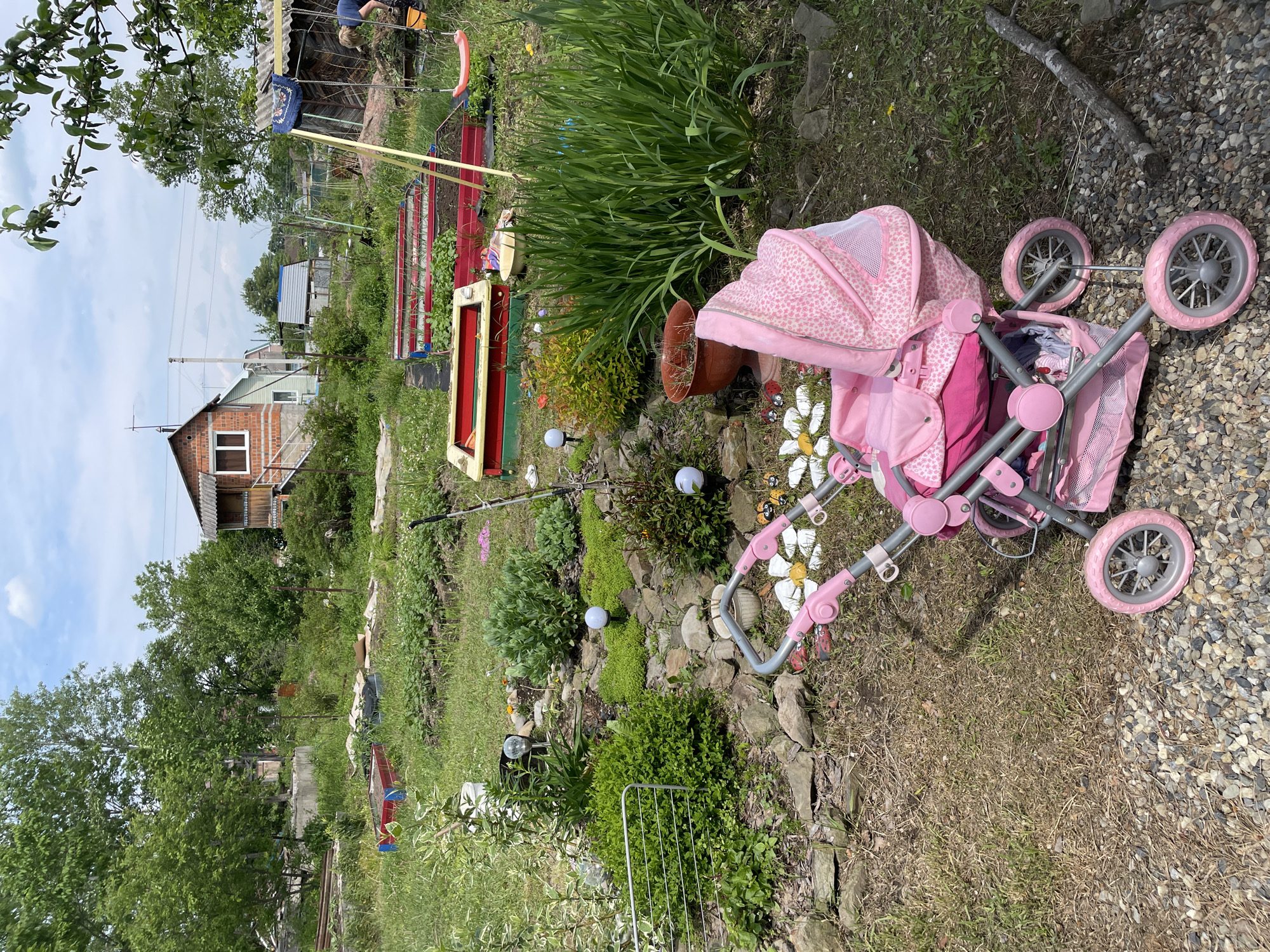 The image depicts a backyard scene in Yasnoe, Russia. In the foreground, a pink toy stroller is partially situated on a gravel path with dry grass. Behind the stroller, there is a garden with various plants, white decorative globes, and small rocks. Further back, there are raised garden beds and a child's swing set. In the background, one can see a brick house and other residential structures amongst trees and vegetation. The sky is partly cloudy. No people are clearly visible in the scene.