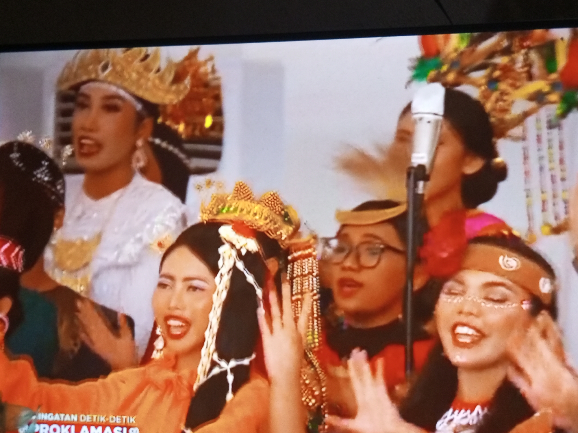 The image displays a group of women in traditional Indonesian attire, likely from Banyudono, Indonesia, adorned with elaborate headdresses and makeup. The women are singing or speaking into a microphone, suggesting a performance or a significant event. The background is blurred but appears to be an indoor setting with decorative elements, possibly a stage or an event hall. The overall mood seems festive and celebratory.