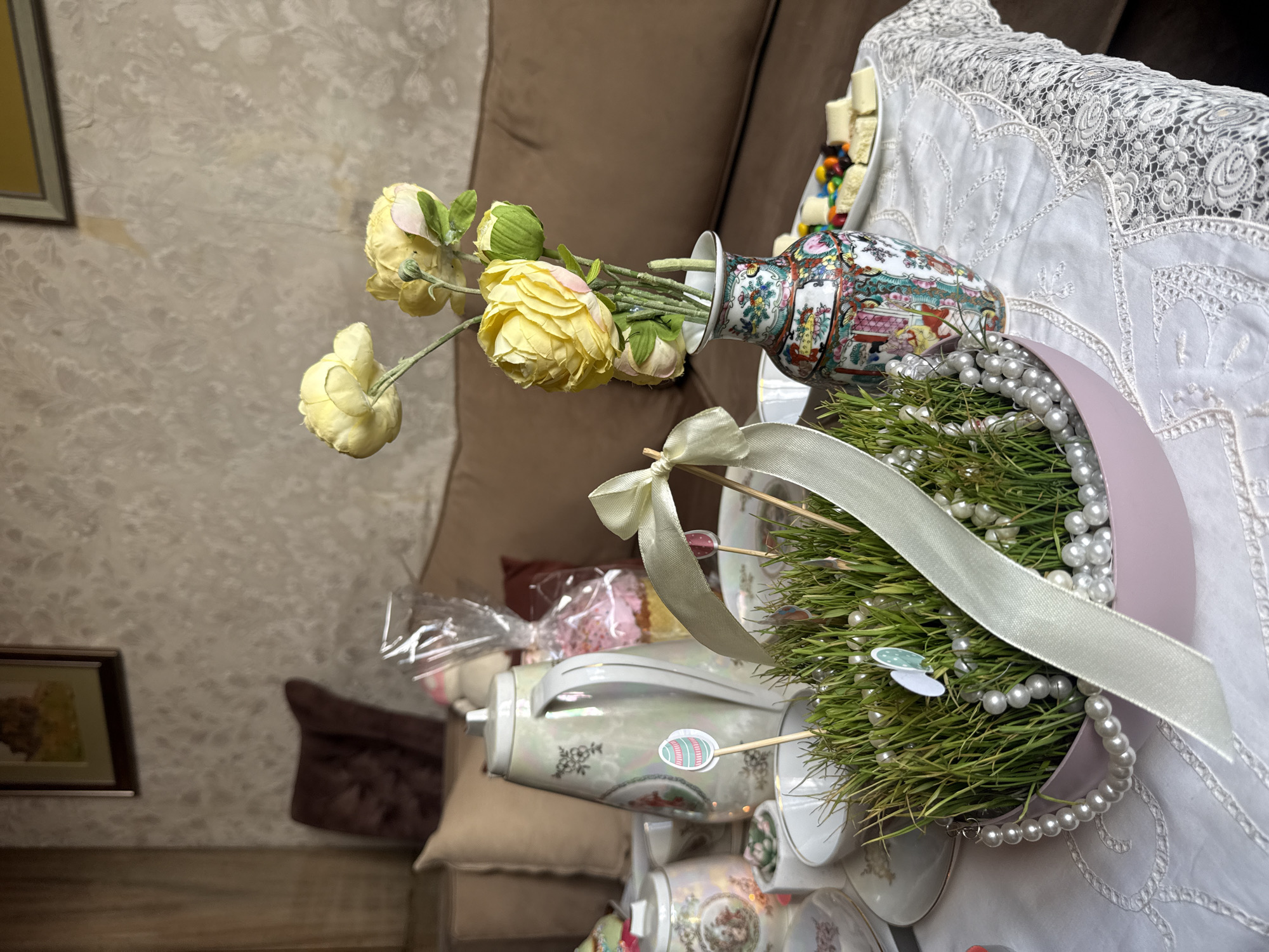 The image showcases a table centerpiece, possibly in a home in Yerevan, Armenia, featuring an elaborate Nowruz display. It includes a vase with yellow flowers, a stack of teacups, and a tray with candies. A decorative arrangement of sprouts, adorned with a white ribbon and a pearl necklace, takes center stage. The table is covered with a white lace tablecloth, and a glimpse of a brown couch and a framed picture suggests a domestic interior setting.