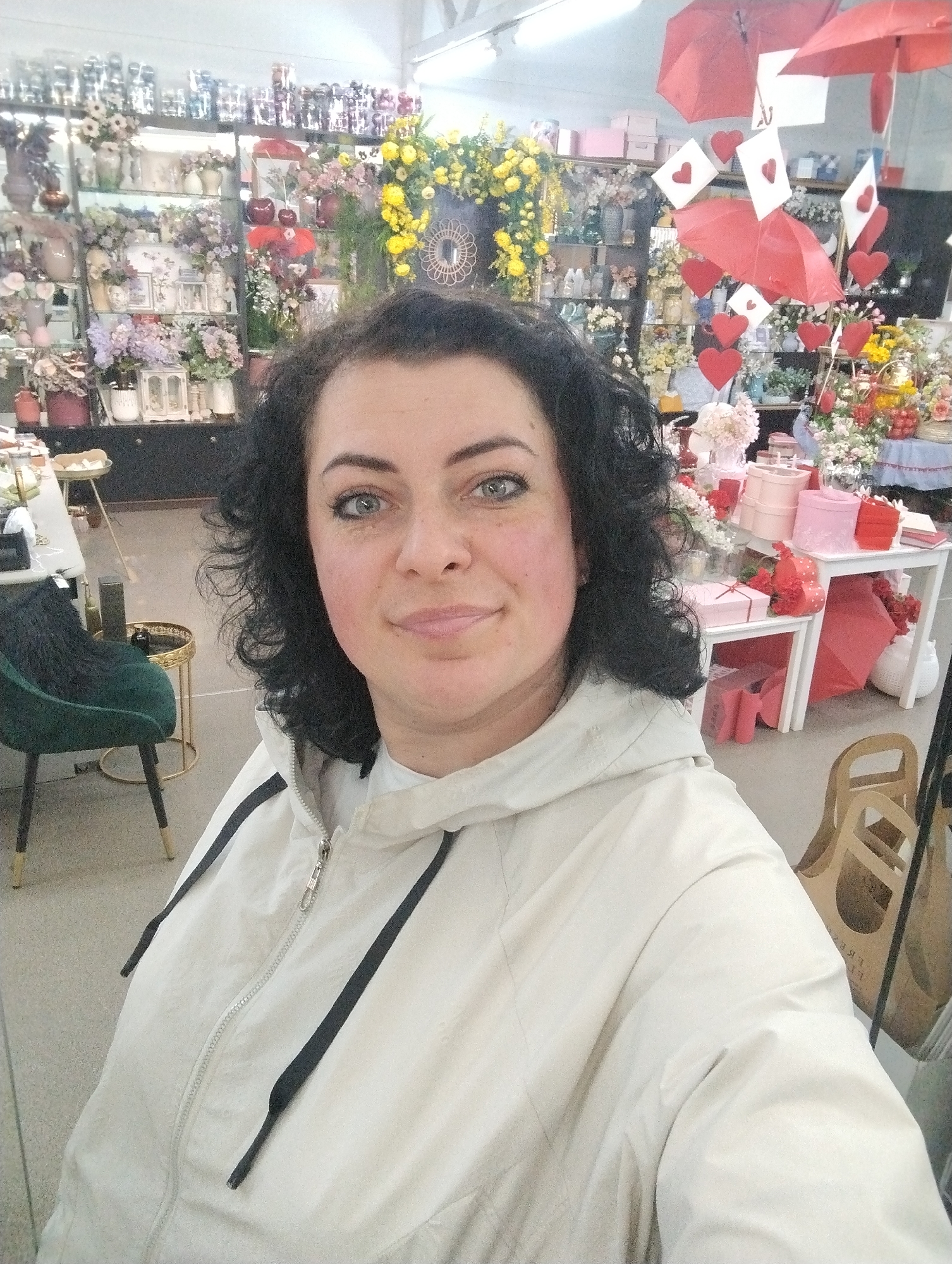 The image shows a woman taking a selfie indoors. She has curly black hair and light eyes, and is wearing a light-colored jacket with black accents. Behind her is a store setting, possibly a flower shop or gift shop, filled with various floral arrangements, decorations, and other items displayed on shelves and tables. Red umbrellas and heart-shaped decorations are visible. The location is likely a retail establishment in Votkinsk, Russia. A green velvet chair is on the left.