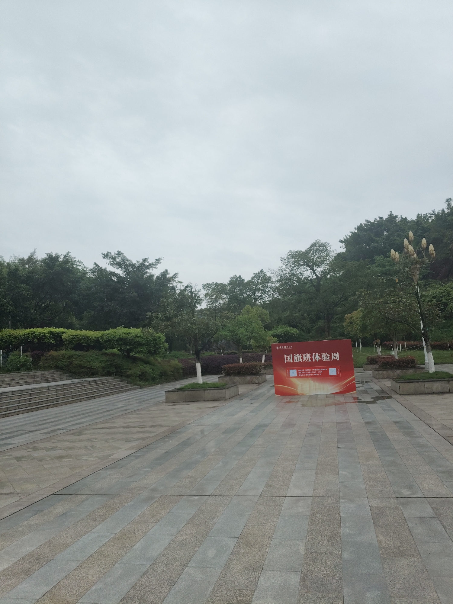 The image shows a public square or plaza in Chongqing High-tech Zone, China, on a cloudy day. The square is paved with grey rectangular stones. A red sign with Chinese characters stands in the middle of the square, possibly advertising an event. Trees and greenery surround the square, and there is a tiered staircase on the left side. A white lamppost with bulbous lights is visible to the right of the sign. The sky is overcast. There are no people visible in the image.