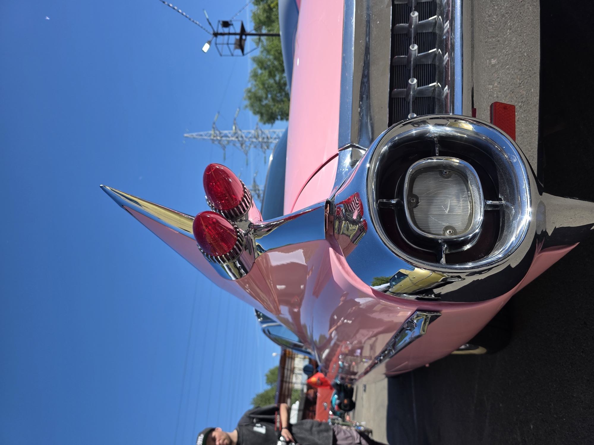 The image shows a close-up of the tail fin and taillight of a pink vintage Cadillac. The car is parked outdoors, likely at a car show or event. In the background, a man wearing a black t-shirt and a baseball cap is visible, standing near other parked vehicles and a red banner. Electrical power lines and towers are also present in the background, suggesting an urban or industrial setting. The sky is clear and blue. The scene is located in Saint Petersburg, Russia.