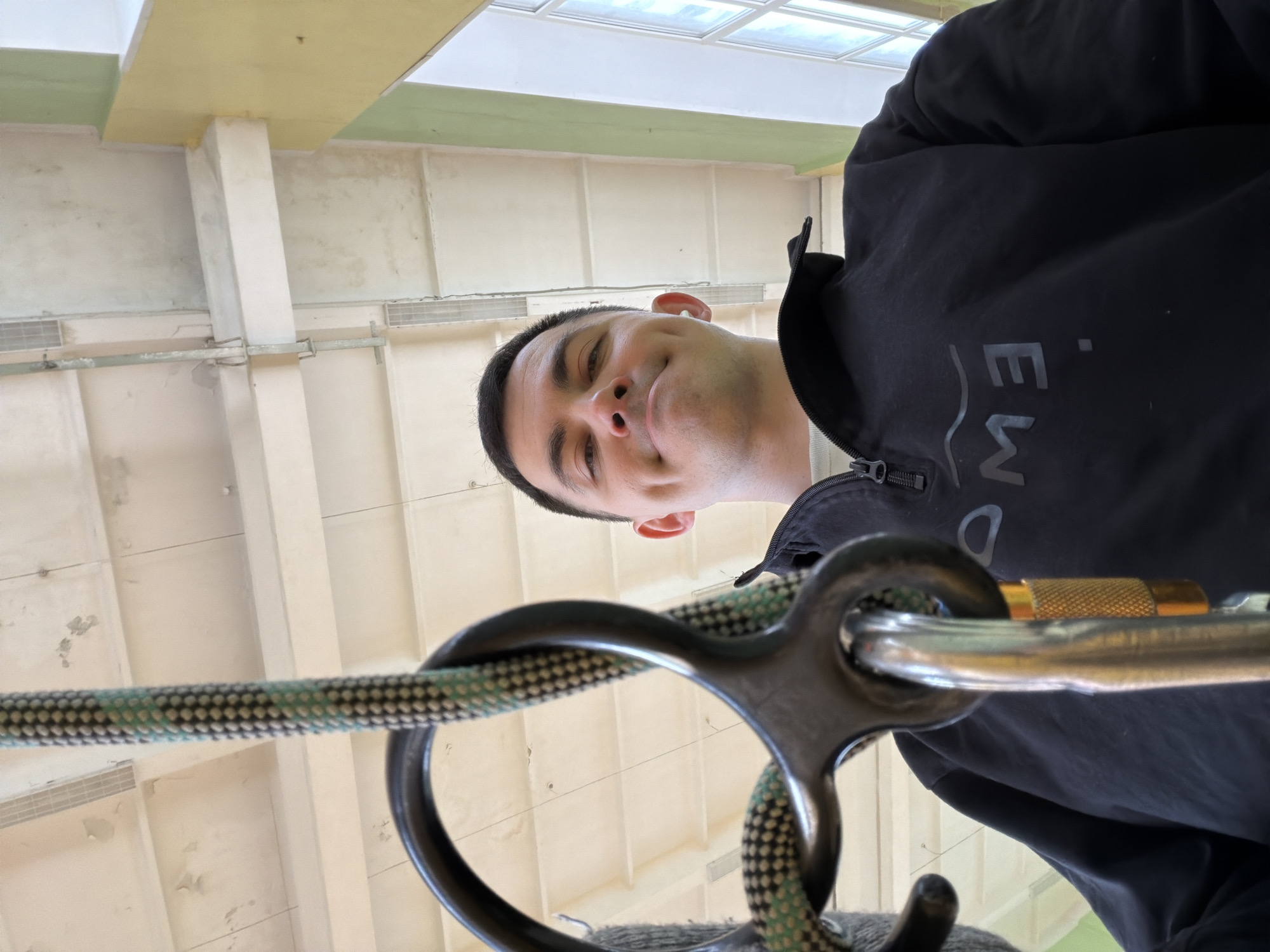 A man with short dark hair and a black jacket is facing the camera. He is using a belay device with a rope in an indoor climbing gym. The location is Izhevsk, Russia. The background features walls and ceilings of the climbing gym.