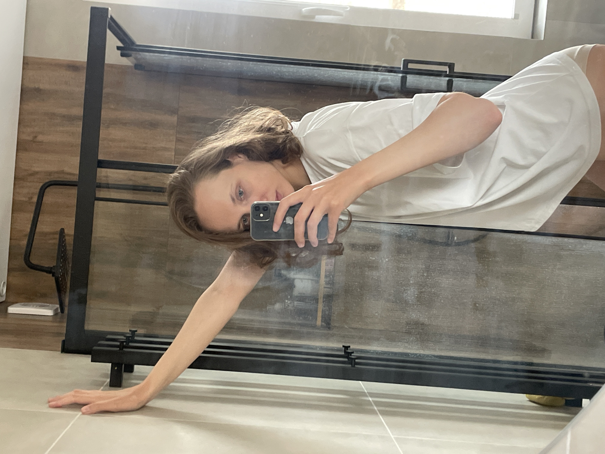 A young woman is captured in a unique selfie, lying down with her head partially supported by a surface and her left arm extended onto a light-colored tiled floor. She holds a dark-colored smartphone in her right hand, pointing it towards herself as she looks directly into the camera with a neutral expression.
She has wavy, light brown hair and is dressed in a loose-fitting white t-shirt and light-colored bottoms. The image appears to be taken through or against a large glass partition or mirror, which creates a partially distorted and reflected view of her body and the surroundings. This glass surface is framed by black metal supports and shows some smudges and reflections.
Behind the glass and the woman, the walls are covered with light brown tiles featuring a wooden texture. A window at the top of the frame allows natural daylight to illuminate the scene, suggesting it is daytime. On the left side, near the floor, there are elements of a black metal structure, a small white rectangular object, and another black metal accessory with a circular, mesh-like part. The setting appears to be an indoor space, possibly a modern bathroom or a room with a large glass screen. There is no visible text in the image.