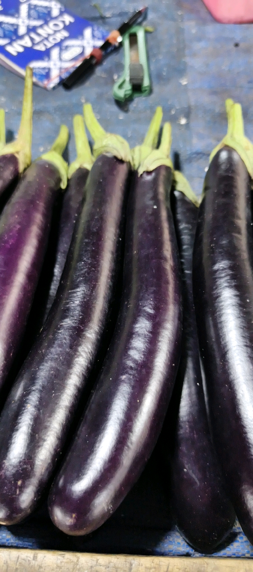 The image shows a close-up, slightly tilted view of a bunch of long, slender eggplants displayed for sale. The eggplants are a deep, glossy purple color with small green stems. They are arranged in a row, with their curved bodies leaning into each other. In the background, a blue and white patterned surface is visible, possibly a table or stall covering. A pen with red and orange accents and a small green cutting tool are also present on this surface, suggesting a market or vendor setting. The overall lighting is dim, highlighting the sheen of the eggplants. It is not possible to determine the specific location within Jambi, Indonesia from this image alone.