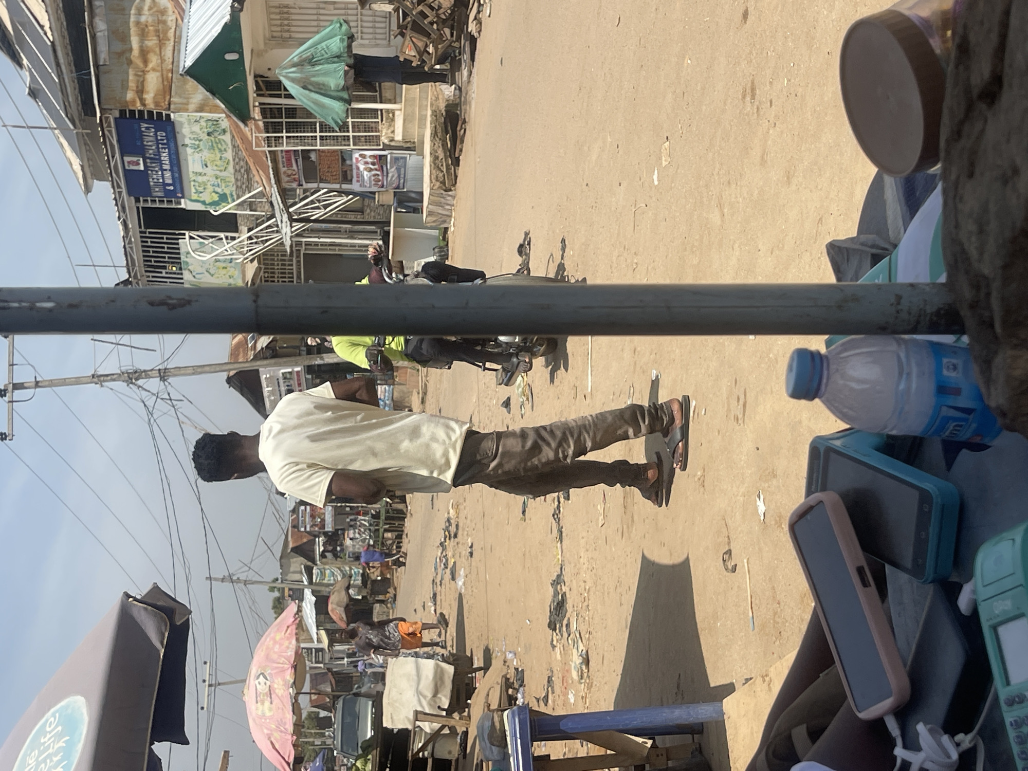 The image depicts a bustling street scene in Abuja, Nigeria, likely in a market area. In the foreground, several mobile phones and a bottle of water are arranged on a surface, suggesting a vendor's stall or a personal setup. A dark-colored metal pole dominates the center of the frame, partially obscuring the view.
In the midground, a man wearing a light-colored t-shirt and dark pants walks away from the camera, his back to the viewer. He is wearing sandals. Further down the street, another person in a lime green shirt is seated on a motorcycle, also facing away. A pink umbrella is visible to the left, and various market stalls and buildings line the street. Debris and sand are scattered across the ground, indicating an unpaved or dusty environment. In the background, a building with a blue sign that reads "WHITEHEART PHARMACY & MINI-MARKET LTD" is visible. The overall atmosphere suggests a lively, everyday commercial activity.
