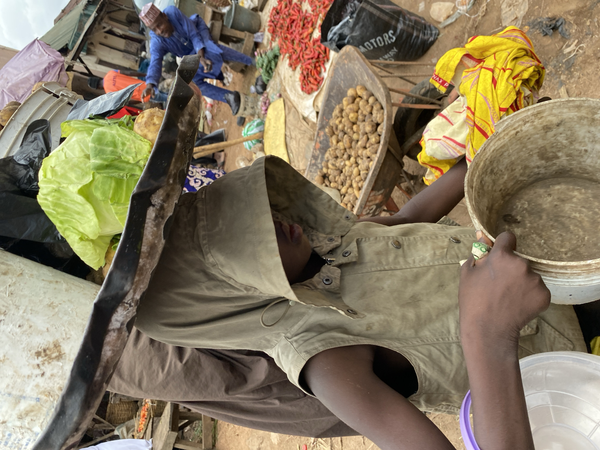 The image shows a person wearing a light brown hooded jacket, with their face obscured. They are holding a light brown bucket in their right hand, which contains a greyish substance. A purple ring with green and white circular patterns is visible on their ring finger. The person is positioned in what appears to be an outdoor market setting.
In the background, there are various goods displayed, including a large head of cabbage, a pile of red chilies, and a wheelbarrow filled with potatoes. A man in a blue traditional outfit and a red cap is visible in the upper left portion of the image, appearing to be engaged in some activity. Several bags, some black with white text reading "MOTORS WAY", and other assorted items are scattered around the scene. A yellow and red patterned cloth is draped over some of the goods. The ground is earthy and appears uneven. The lighting suggests it is daytime, likely overcast given the soft shadows. The overall scene suggests a busy marketplace, possibly in Bukuru, Nigeria, with a focus on fresh produce.