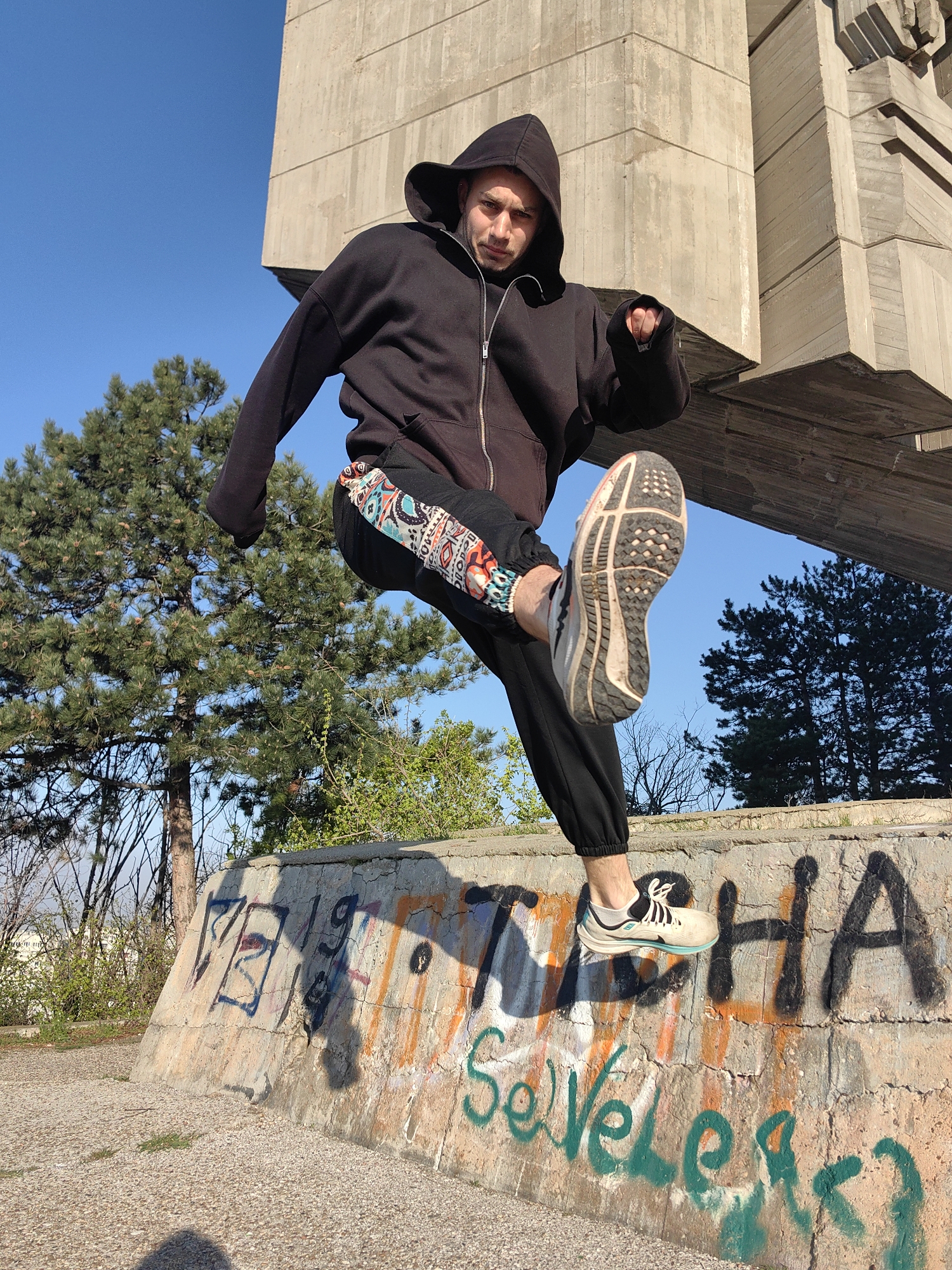 A man in a black hoodie and sweatpants with patterned trim on the sides is captured mid-air, performing a high kick. He's wearing athletic shoes. The photo is taken outdoors, possibly in a park setting. Behind him are trees and a large, abstract concrete structure. The man appears to be standing on a low concrete wall covered in graffiti; the word "NIKA" is partially visible. Based on the style of the concrete structure, the graffiti, and the park-like setting, the location is most likely the Pantheon in Varna, Bulgaria. The day appears sunny with a blue sky.