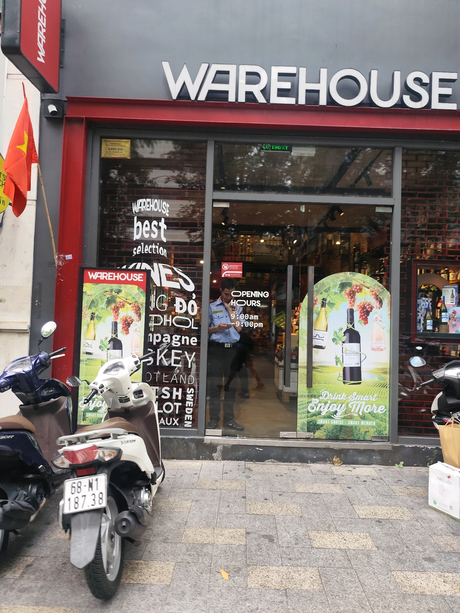 The image shows the exterior of a "WAREHOUSE" store located in Ho Chi Minh City, Vietnam. A large white sign with "WAREHOUSE" is mounted above the entrance. Two motorcycles are parked on the sidewalk in front of the store. One is blue and parked on the left, and the other is white and parked on the right, closer to the entrance. The white motorcycle has a license plate with the number "68-M1 187.38". A Vietnamese flag is visible on the left side of the image, attached to a pole. The store has a glass entrance with signs on it indicating "WAREHOUSE best selection" and "OPENING HOURS 9:00 am - 9:00 pm". A security guard in a blue uniform is standing near the entrance, looking at his phone. Inside the store, shelves stocked with bottles are visible. A poster displaying grapes and wine bottles is placed near the entrance, with the text "Drink Smart Enjoy More". The ground in front of the store is paved with stone tiles.