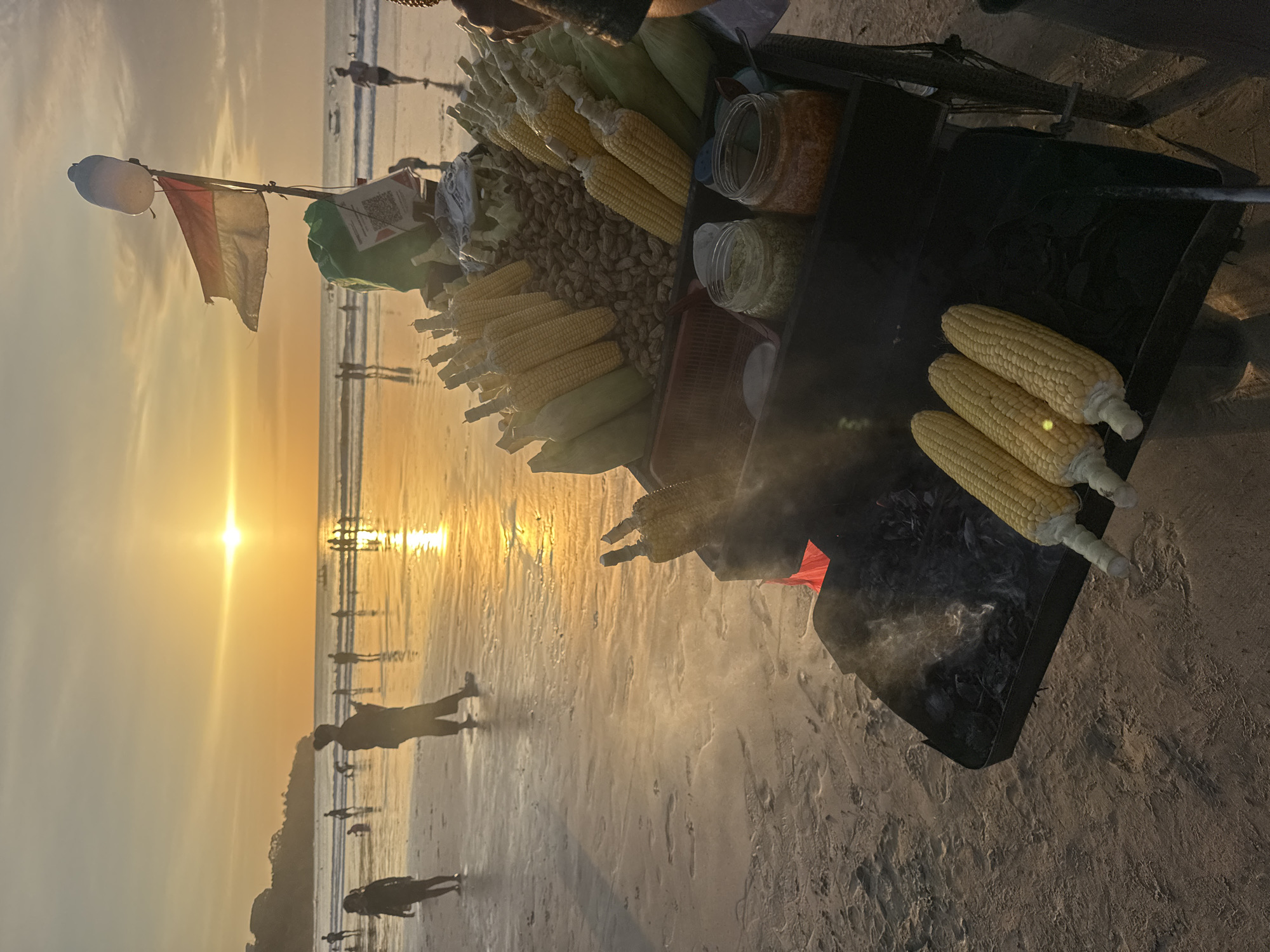 In Kuta Selatan, Indonesia, a beachside vendor grills corn on the cob over hot coals at sunset. The golden sun casts a warm glow on the wet sand, reflecting the sky and silhouetting people walking along the shore. A cart laden with unhusked corn, peanuts, and condiments sits in the foreground, with the Indonesian flag fluttering above. Smoke rises from the grill, mingling with the humid ocean air as a gentle breeze rustles the leaves of nearby trees. The scene is serene, with the sounds of waves crashing and distant chatter creating a tranquil atmosphere.