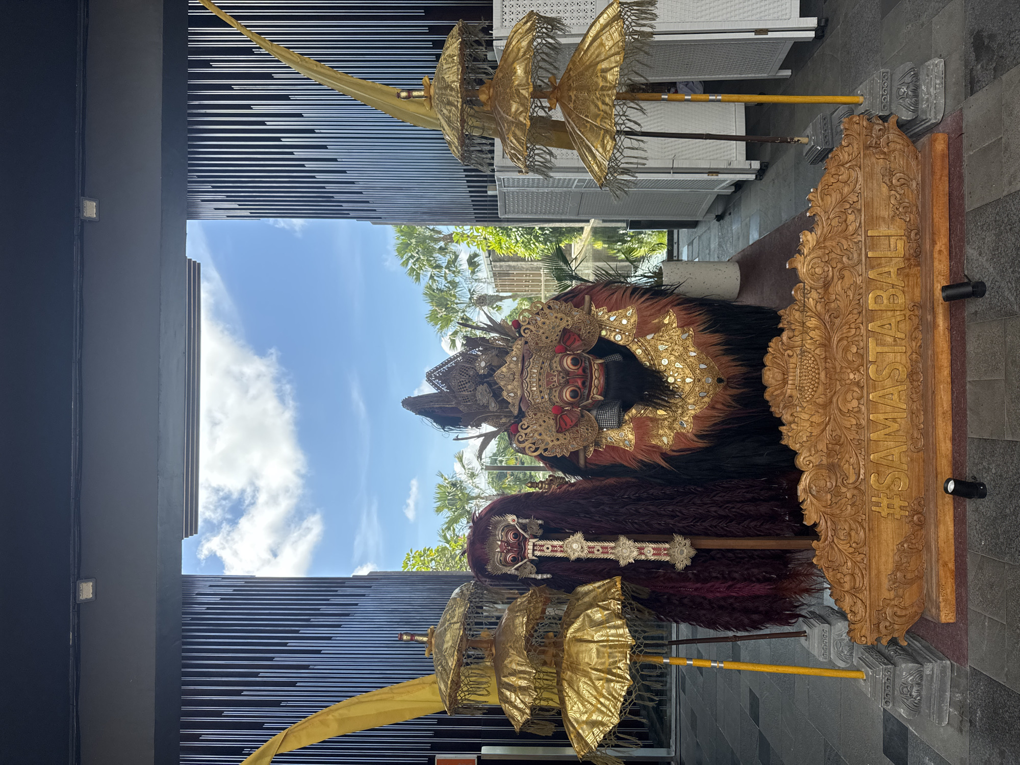 The image shows two traditional Balinese Barong dance costumes displayed outside, likely in Kuta Selatan, Indonesia. The costumes, richly decorated with gold and brown fur-like materials, are positioned next to a carved wooden sign with the text "#SAMASTABALI" vertically oriented on it. Several golden umbrellas are placed near the costumes. The backdrop includes a modern building with horizontal slats and a view of a blue sky with scattered clouds and some palm trees. The ground is tiled.