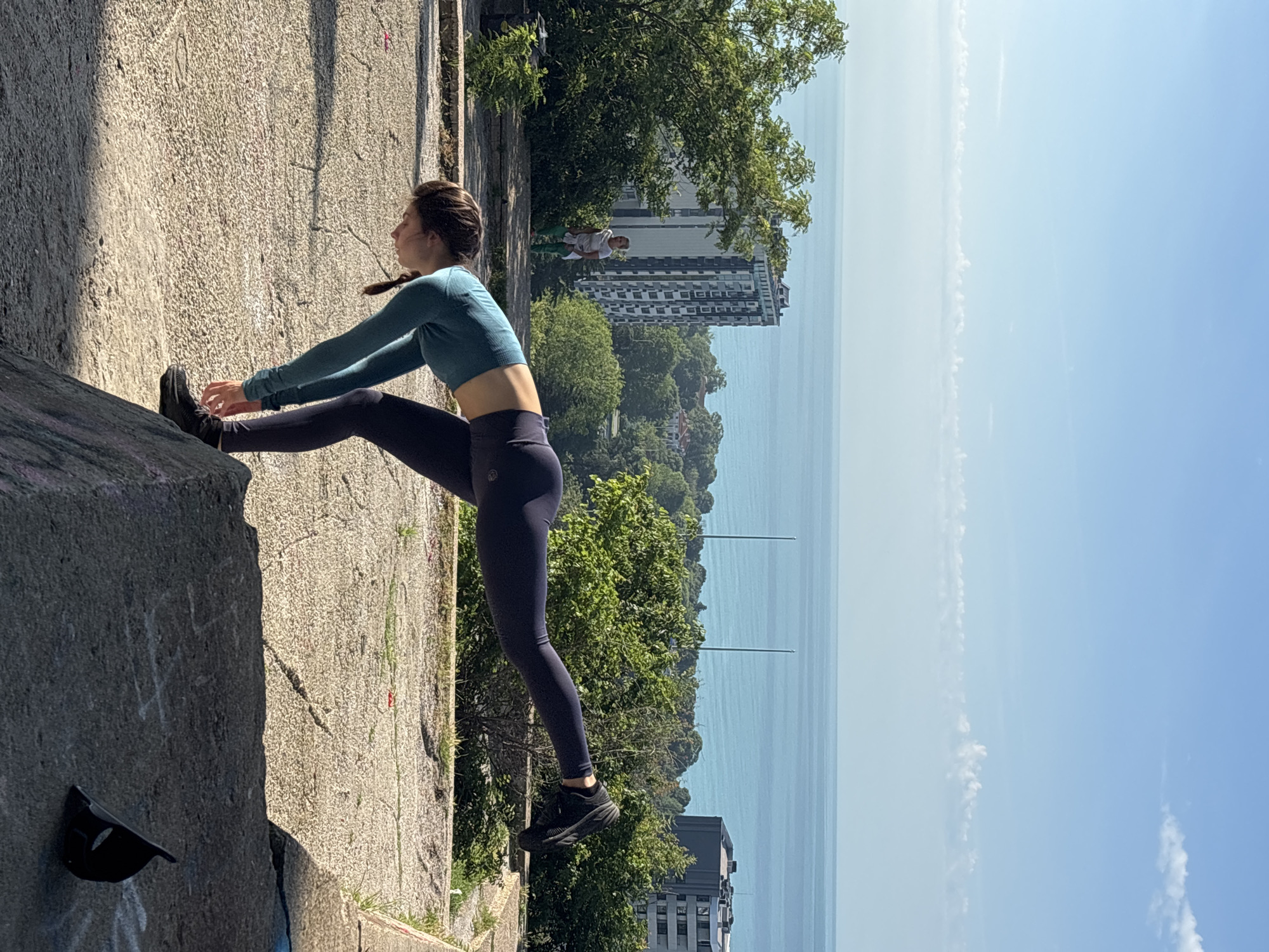 A young woman in athletic wear is captured mid-stretch in an outdoor setting. She is wearing a long-sleeved teal crop top and dark blue or black leggings, with black athletic shoes. Her hair is pulled back, possibly in a braid. She is bending forward, holding her raised front foot, indicating an exercise or stretching routine. A faint logo is visible on her left hip, possibly on her leggings.
The scene features a rugged, light-colored concrete or rocky ground in the foreground, with strong sunlight casting shadows. In the middle ground, there are green trees, and beyond them, several modern-looking buildings. Further in the distance, a vast expanse of calm, blue sea or ocean is visible under a clear, bright blue sky with a few wispy clouds. Another person, dressed in a white t-shirt and light-colored pants, is standing in the background among the trees. A black cap is resting on a lower concrete ledge in the bottom left corner of the frame.
The bright lighting and clear sky suggest it is a sunny day, likely mid-day. The presence of the sea and urban structures aligns with the given context of Varna, Bulgaria, a coastal city. No discernible legible text is visible other than the small logo on the woman's attire.