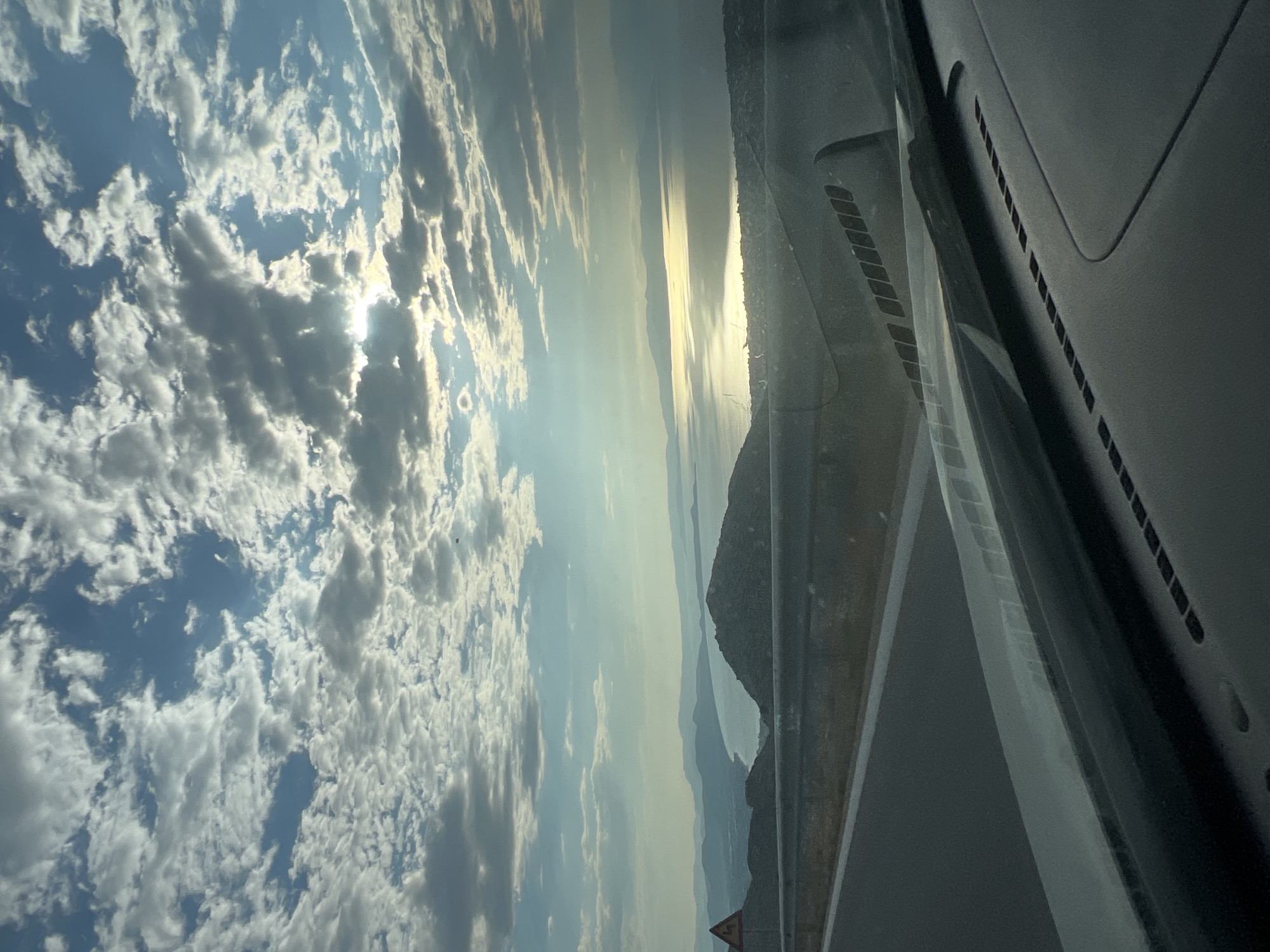 The image is a vertical, eye-level, wide shot captured from inside a vehicle looking out at a scenic coastal view under a cloudy sky. In the foreground, the dark dashboard of the vehicle is visible, with the windshield partially obscuring the view. A road curves to the left, with a guardrail separating it from a steep drop-off.
Beyond the road, a dark, hilly landscape stretches out, leading to a body of water that reflects the bright, sunlit clouds. The sun is partially obscured by clouds, creating a shimmering effect on the water. In the distance, layers of hazy mountains recede into the horizon. A small, dark object, possibly a bird or drone, is visible in the sky above the distant mountains. A yellow triangular warning sign with a black zigzag symbol is present on the left side of the road.
The scene appears to be located in or near Nea Makri, a municipal unit within the Marathon municipality of Attica, Greece, known for its coastal beauty and hilly terrain. The overall atmosphere is serene and expansive, highlighting the natural landscape under a dynamic sky. There are no people visible in the image.