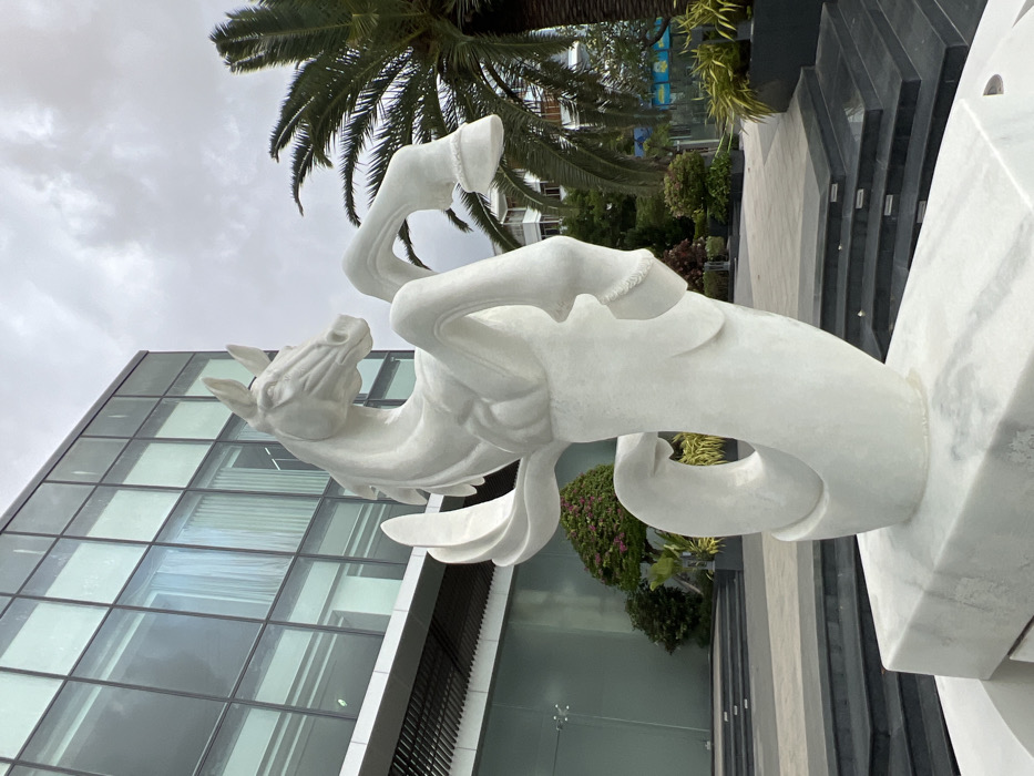 An outdoor scene in Nha Trang, Vietnam, features a large, white marble sculpture of a horse rearing, its forelegs elevated and head turned right, supported by a stylized wave-like base. The sculpture is mounted on a multi-tiered white marble pedestal. In the background, a modern building with a reflective glass facade occupies the left, partially reflecting the sky. To the right of the sculpture, a tall palm tree with green fronds stands prominently. The ground level visible includes dark grey paved steps and planters containing varied tropical foliage, including a bush with pink blossoms. The sky is uniformly overcast. No individuals or discernible activities are present.