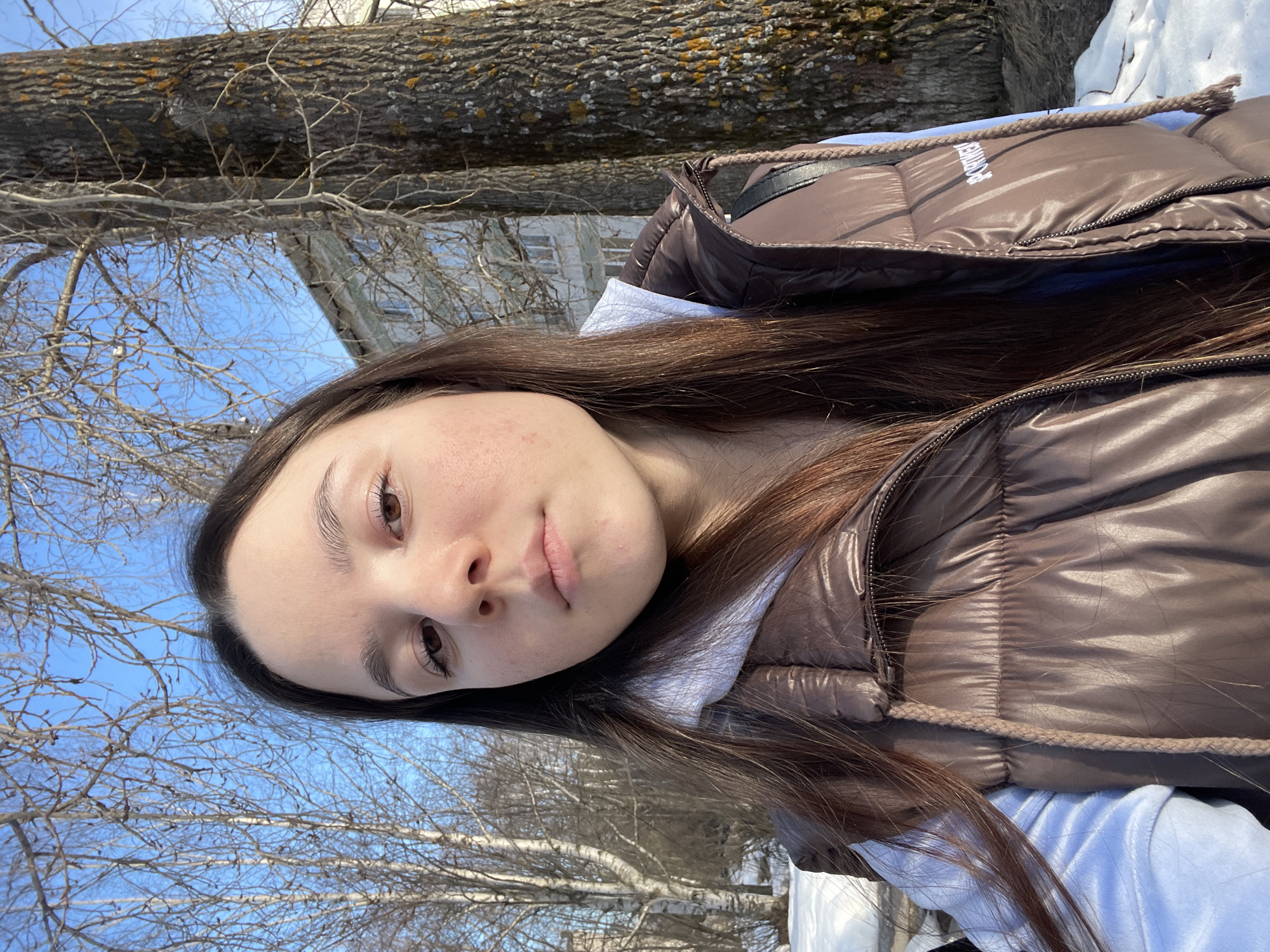 A young woman with long brown hair, wearing a brown puffy vest over a light blue hoodie, is posing for a selfie outdoors. Her face is slightly tilted towards the camera. The background includes bare trees and a clear blue sky. A building is visible in the upper left corner. Based on the presence of birch trees and the architecture, it is most likely a location within the city of Chaikovsky, Russia. The ground appears to have some snow patches.