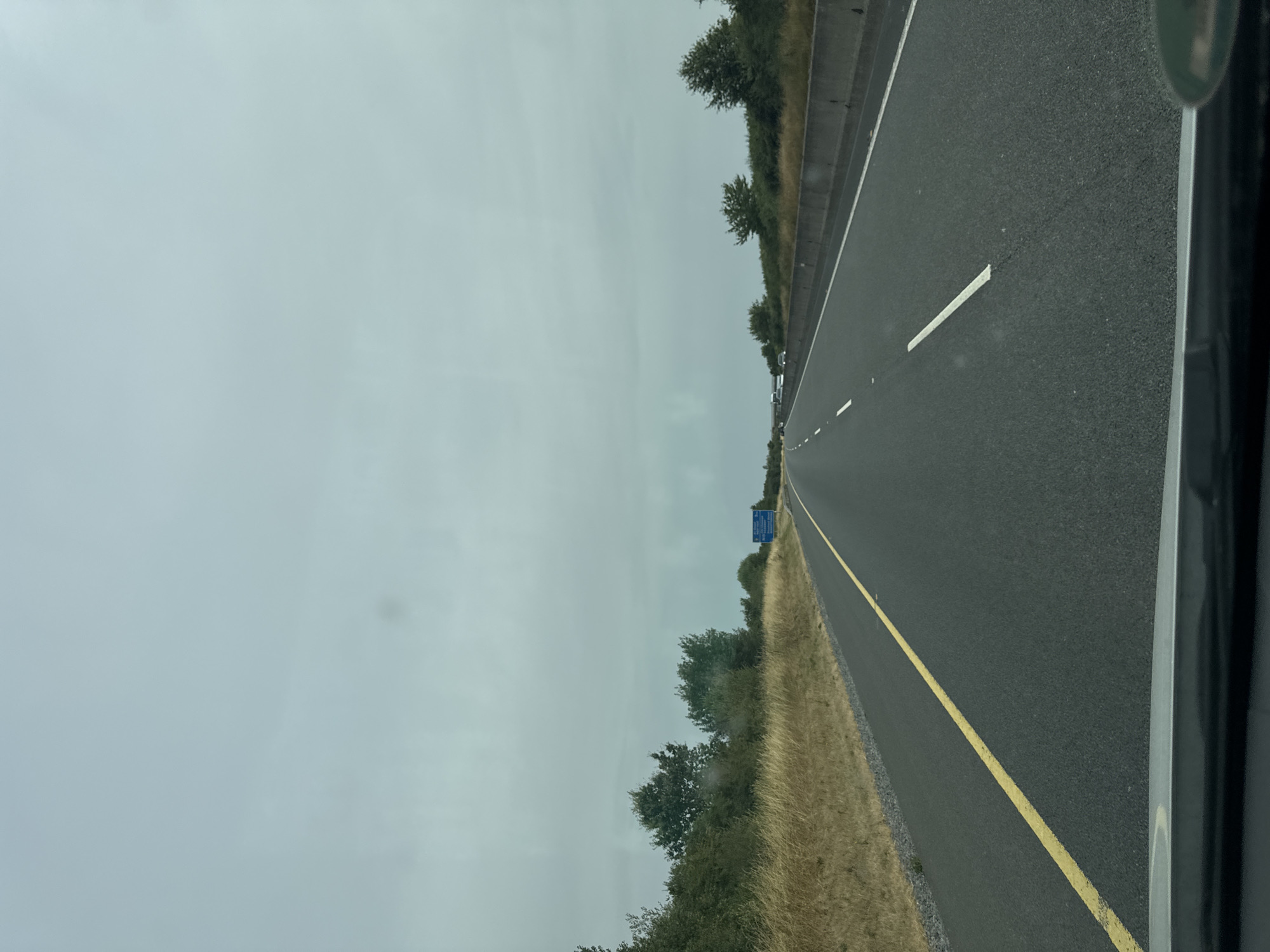 The image shows a view from inside a vehicle traveling on a highway in Ireland. The highway is paved with asphalt and has a yellow line separating the lanes. The sky is overcast and the landscape consists of dry grass and trees on either side of the road. There is a blue highway sign visible in the distance to the right. There are no people or other vehicles immediately visible in the foreground. The location is not specifically identifiable as Rivergrove, Ireland from the image alone.