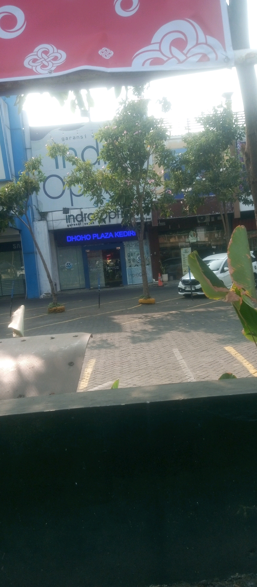 The image shows a street scene in front of a building that appears to be a commercial establishment in Kediri City, Indonesia. A red banner with white circular and knot-like designs is hung above. Below the banner, a storefront is visible with a blue neon sign that reads "DHOHO PLAZA KEDIRI". Trees with green foliage and some small pink flowers are planted in front of the storefront. The ground is paved with cobblestones, and there are yellow parking lines visible. A white car is parked on the right side of the image. The overall scene is well-lit by sunlight, suggesting it is daytime.
