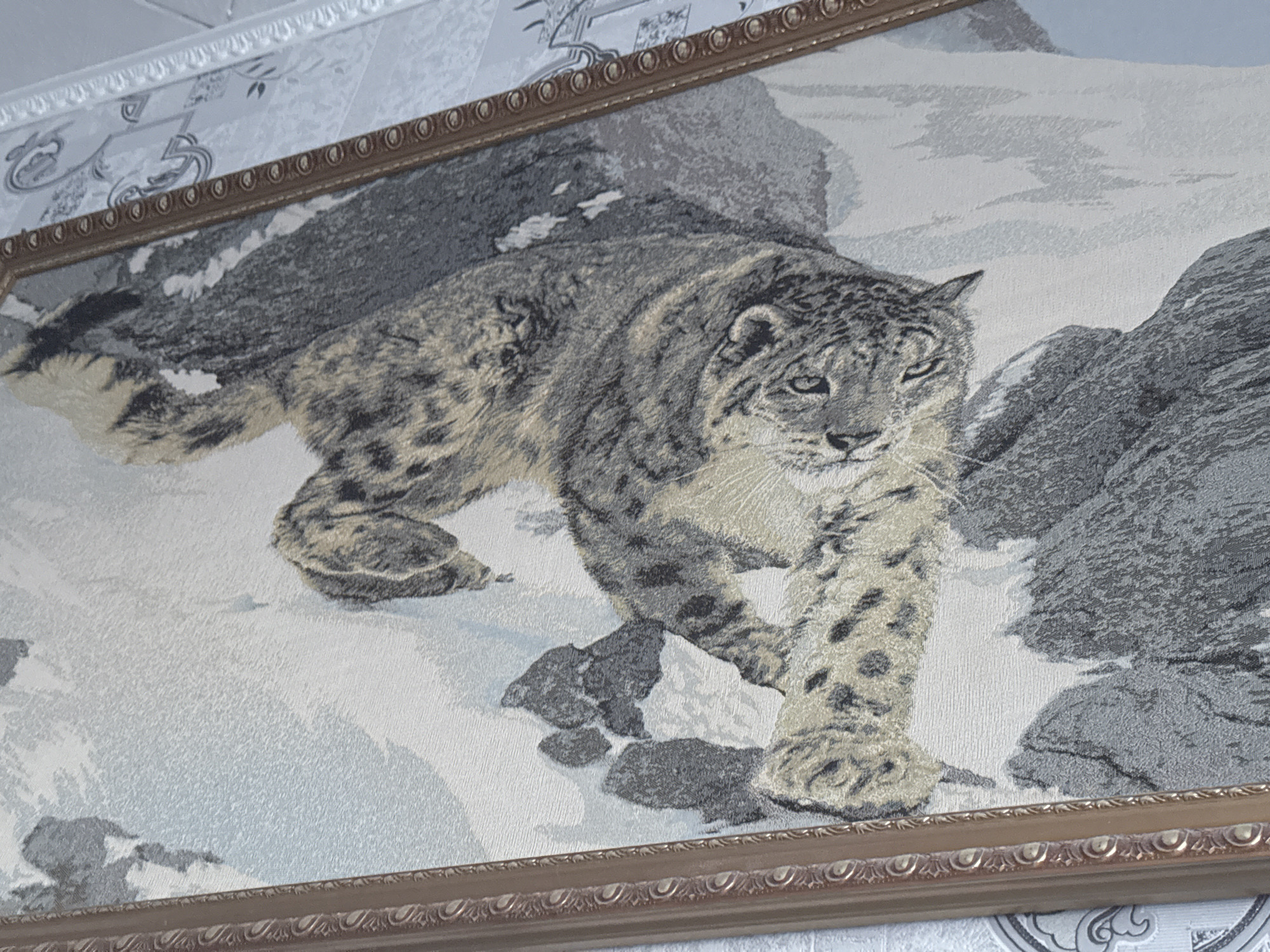 A framed tapestry of a snow leopard walking on snow-covered rocks hangs on a wall with patterned wallpaper. The snow leopard is depicted in shades of gray, black, and tan, with its distinctive spotted fur and piercing green eyes. The background features a rocky, mountainous landscape with patches of snow. The tapestry has a decorative wooden frame with an ornate border. The wallpaper behind the tapestry is light in color with a subtle floral or damask pattern. A textured white ceiling molding is visible above the tapestry.