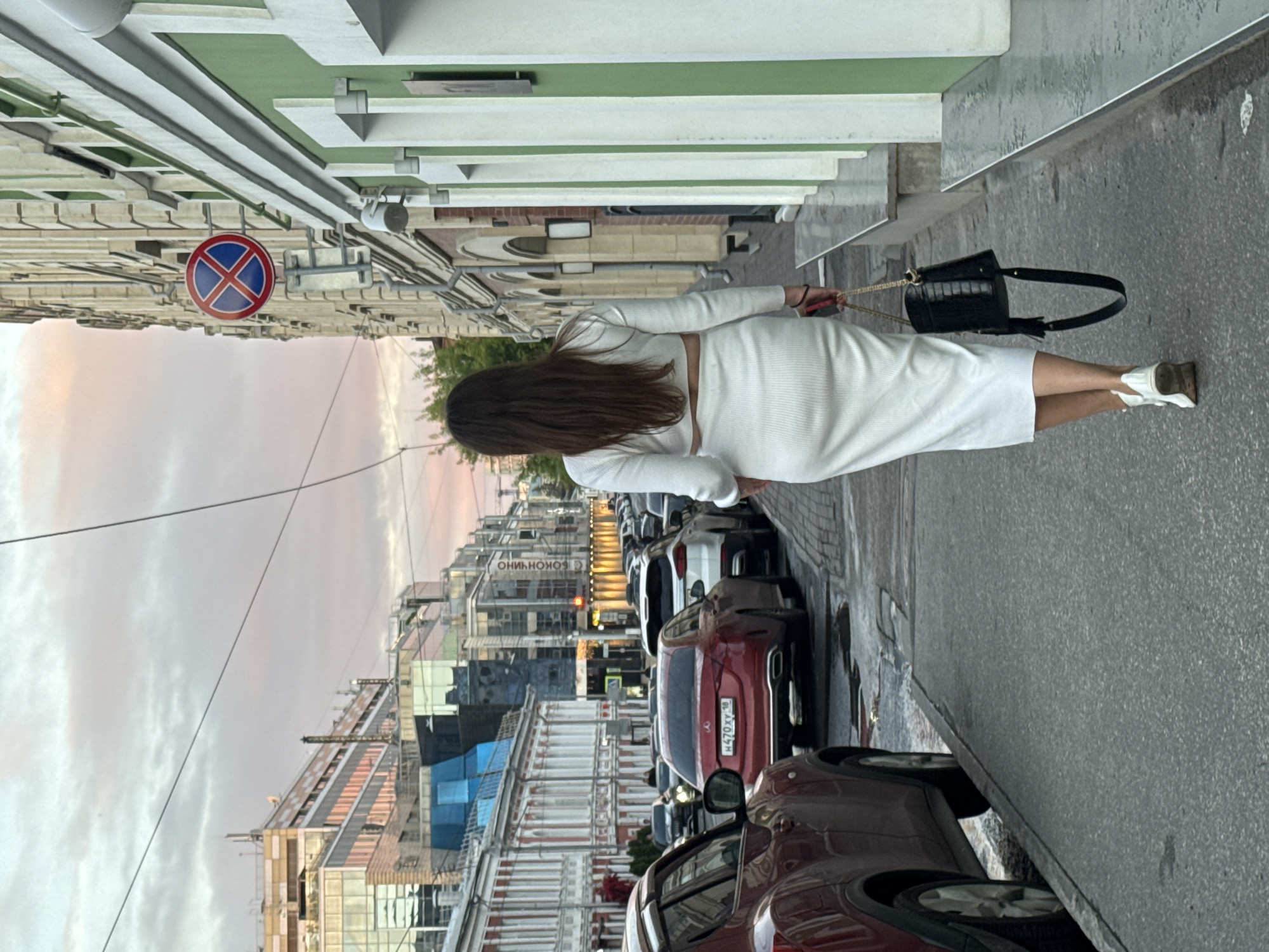 A woman walks down a city street in Nizhny Novgorod, Russia, on a cloudy day. The woman, seen from behind, wears a white, form-fitting, two-piece outfit consisting of a long-sleeved crop top and a midi skirt. She carries a black handbag with gold accents and is wearing white sneakers. The street is lined with buildings, including a red and white building on the left and a green and cream building on the right. Several cars are parked along the street. A "no parking" sign is visible on the building to the right. The sky is overcast with hints of pink from the sunset.