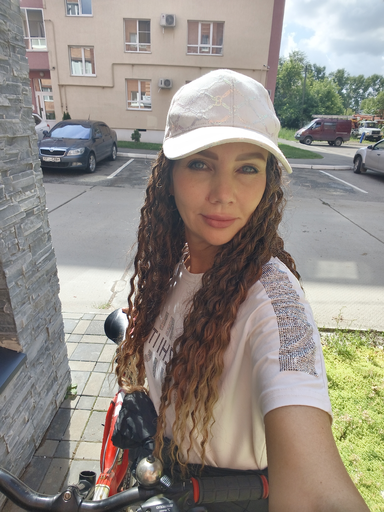 An outdoor, eye-level, medium-shot selfie shows a woman with long, curly brown hair and blue eyes wearing a white baseball cap and a white t-shirt with a sparkly design on the sleeve and chest. The woman is looking at the camera with a slight smile. She is standing next to a bicycle. The background shows a brick building with windows, parked cars, and trees. The lighting suggests it is daytime and sunny. The location is identified as Unknown, Unknown.