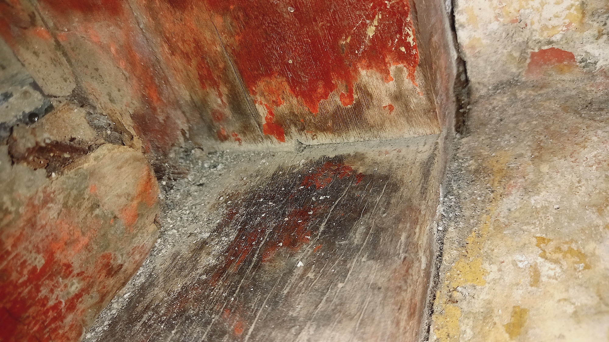 The image shows a close-up view of weathered and damaged corner of a structure, possibly a wall or a foundation, in Leuwisadeng, Indonesia. The surfaces are rough and show signs of significant wear and tear.
One surface, appearing to be wood or a similar material, is covered in peeling red paint. The wood grain is visible underneath the paint, and there are streaks and patches where the paint has worn away, revealing the lighter wood. This surface is oriented diagonally across the frame, suggesting it is part of a corner.
Adjacent to this, another surface is visible. It is rough and textured, appearing to be made of concrete or plaster. This surface is a lighter, sandy color with patches of yellow and pinkish-red. It also shows signs of abrasion and damage.
The corner where these two surfaces meet is filled with small, loose debris, resembling gravel or sand. This debris is a mix of gray, white, and brown particles, with some faint reddish specks. The overall impression is one of neglect and decay.
There are no people, animals, or discernible objects other than the structural elements themselves. The lighting is dim, casting subtle shadows that accentuate the textures and imperfections of the surfaces. The environment appears to be outdoors, given the weathering and the natural materials involved, but the specific context within Leuwisadeng, Indonesia, is not directly evident from the image alone, beyond the provided location information.