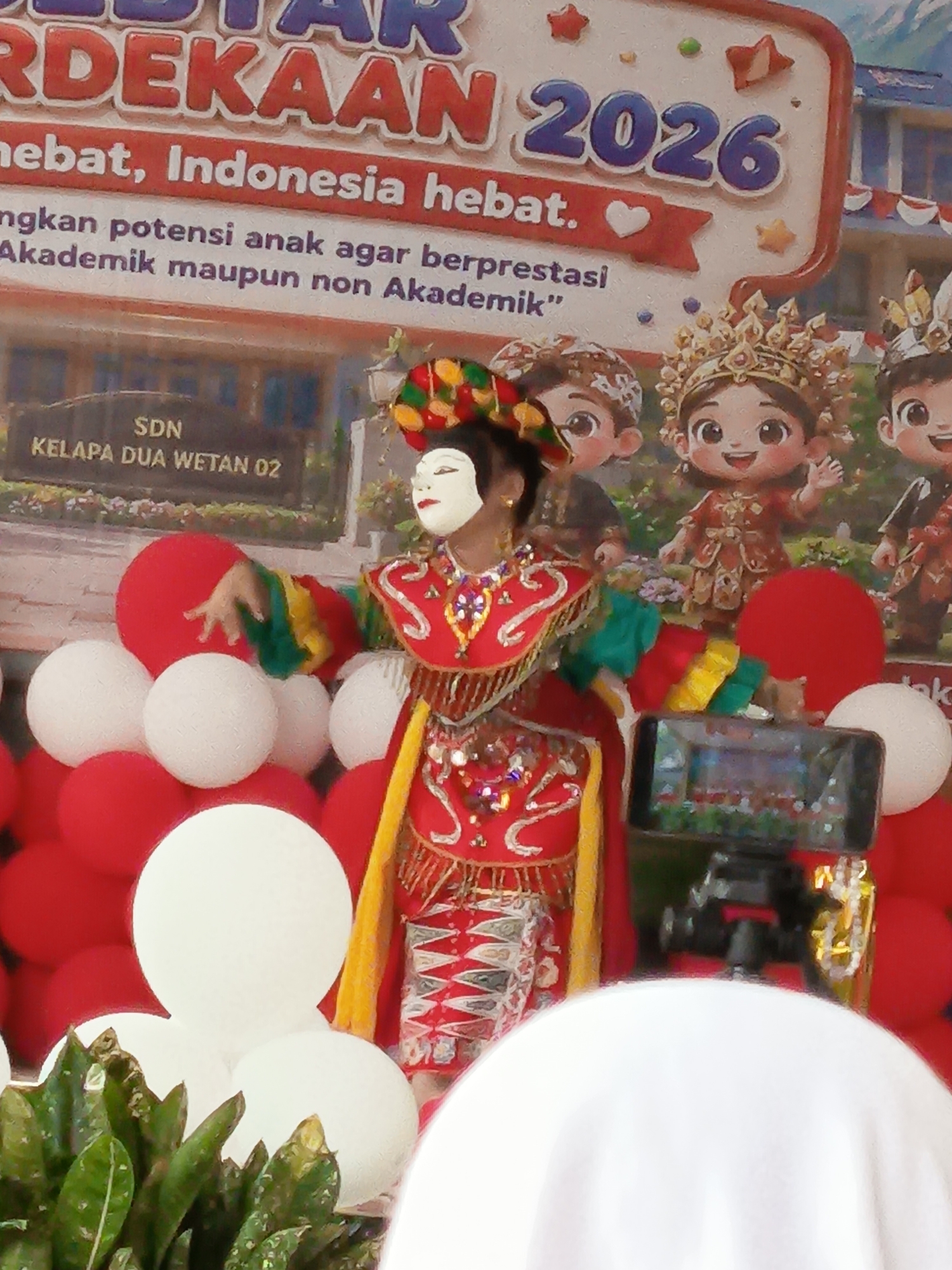 The image shows a young person in traditional Javanese costume, complete with a mask and elaborate headdress, performing a dance on a stage. The stage is decorated with red and white balloons. Behind the dancer is a banner with text that appears to be in Indonesian, including "SDN KELAPA DUA WETAN 02," which indicates the location is the Kelapa Dua Wetan 02 State Elementary School. The banner also displays "MERDEKA 2026" and other text that seems to be motivational. Several animated cartoon characters in traditional Indonesian attire are depicted on the banner. A smartphone on a tripod is positioned in front of the stage, seemingly recording the performance. In the foreground, there are several white and red balloons, and some green foliage. The overall scene suggests a cultural event or celebration at a school in East Jakarta, Indonesia.