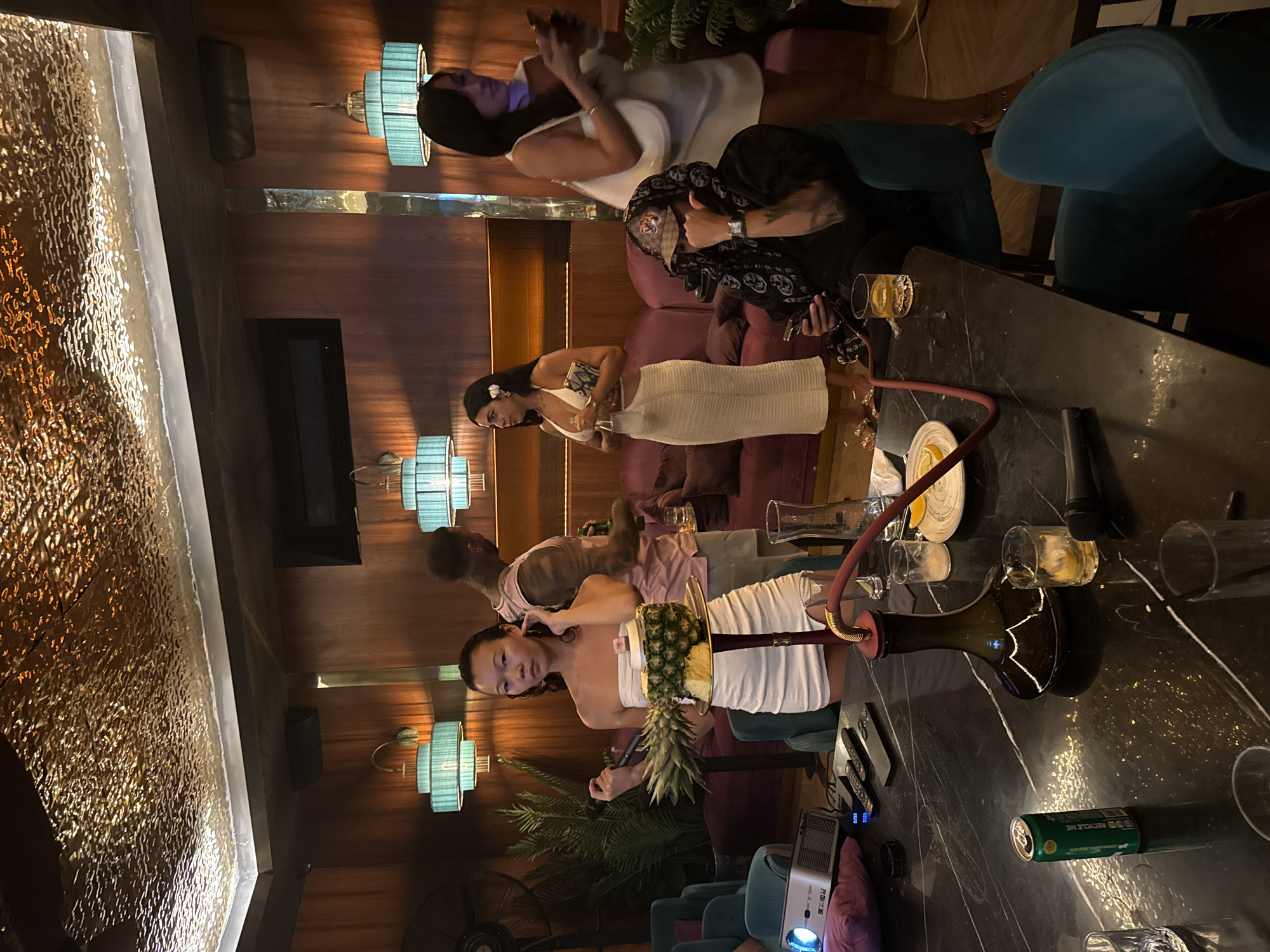 The image displays an indoor evening scene within a dimly lit, upscale lounge or bar, likely located in North Kuta, Indonesia. The atmosphere is relaxed and social, with several individuals gathered.
In the mid-ground, a woman wearing a white strapless top and skirt is seated at a dark, marble-patterned table, looking towards the viewer. A unique shisha pipe, prominently featuring a large, fresh pineapple as its head, stands on the table beside her. Another black shisha pipe is also visible on the table, along with several glasses containing various beverages, some with ice and lemon. Remote controls, a small projector, and a green can with "RECYCLE ME" printed on it are also on the table.
Behind this central figure, another woman in a white dress with a flower in her hair is standing, holding a drink. Further back, a man in a light pink shirt stands partially visible. On the left side of the frame, a woman in a white dress is seated on a plush maroon sofa, looking at her phone. Another person in a black patterned hoodie and cap is seated further right, partially obscured.
The lounge is adorned with decorative elements, including warm ambient lighting from teal-colored wall sconces with layered shades. The walls are paneled in wood, complementing the comfortable seating arrangements, which feature teal armchairs and maroon sofas. A striking, large shimmering golden textured wall or water feature runs along the left side of the space, adding a luxurious touch. Potted green plants are strategically placed throughout the area, and a black fan is visible in the lower left corner. The overall impression is one of a sophisticated social gathering.