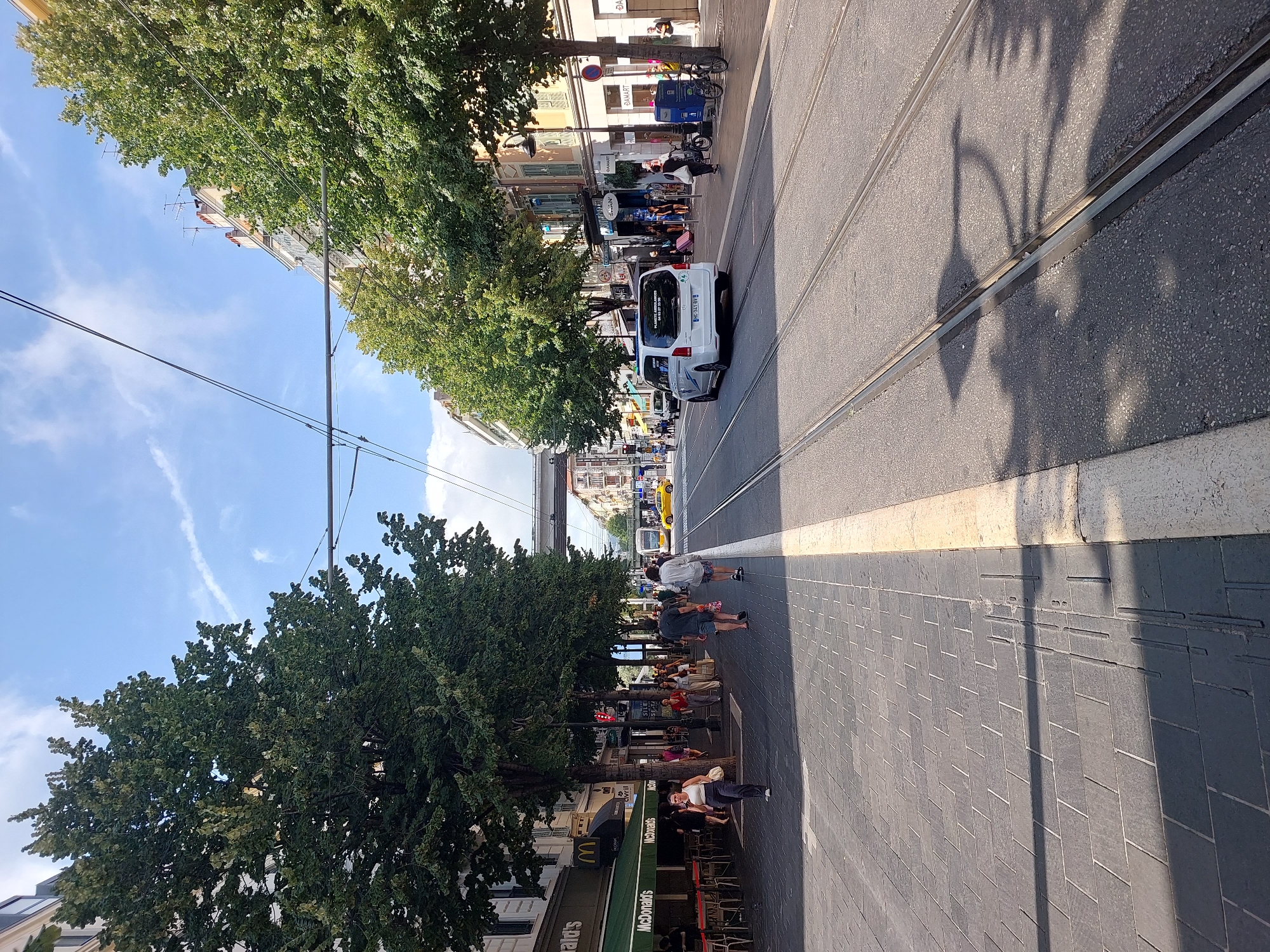 This is a street scene in Nice, France, specifically on Jean-Médecin Avenue, which features tram tracks. On the left side of the image, a McDonald's restaurant is visible with outdoor seating. A tree-lined sidewalk runs parallel to the street, occupied by pedestrians walking in both directions. To the right of the sidewalk, tram tracks are embedded in the asphalt road. Several vehicles, including a white van and a yellow tram, are present on the road. Buildings line the opposite side of the street, and overhead wires for the tram are visible. The sky is blue with some scattered clouds.