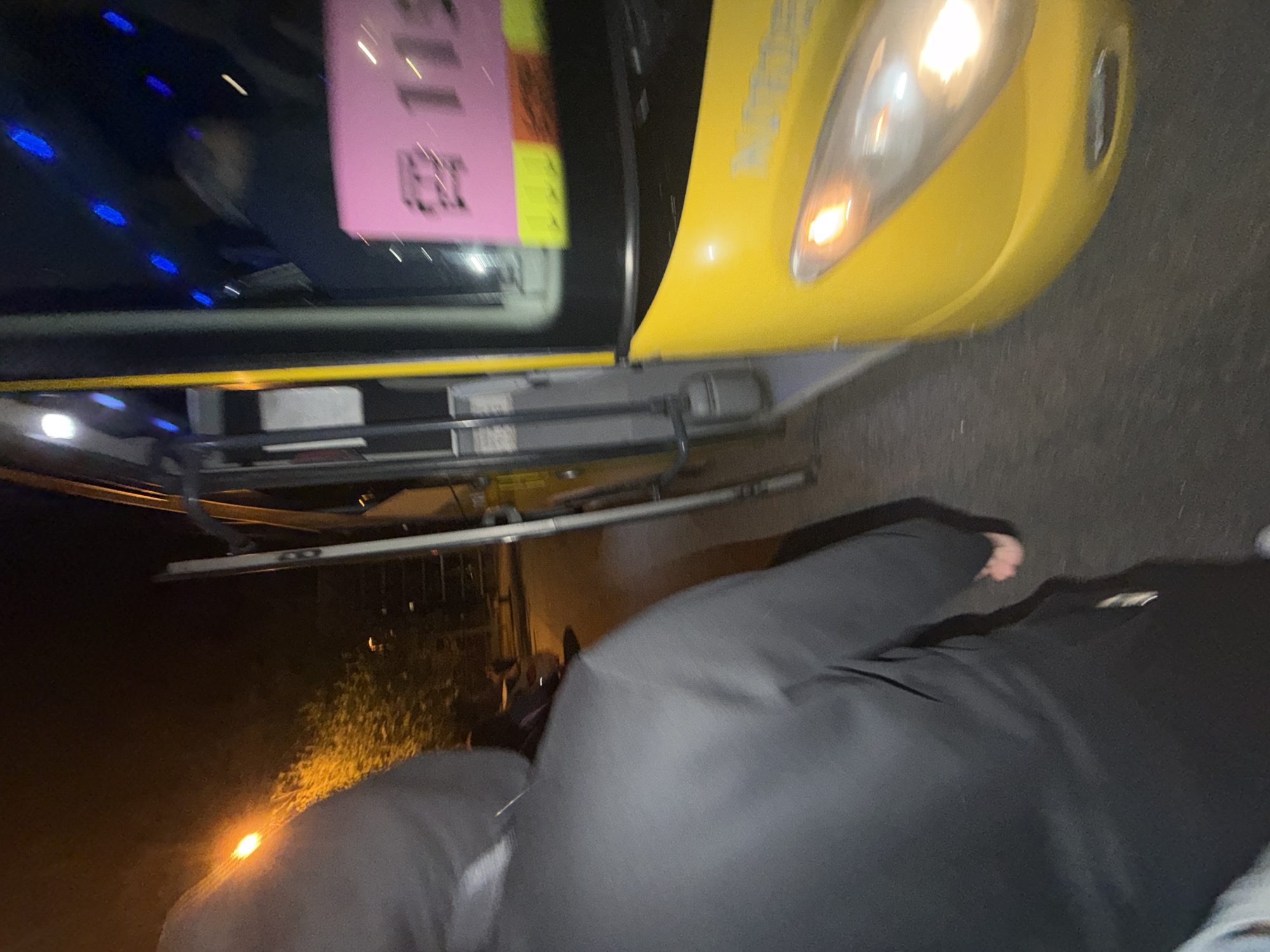 The image shows a person standing in front of a yellow taxi, possibly in Vila Real, Portugal. The taxi has a pink paper sign on the driver's side window. The scene appears to be taken at night due to the poor lighting. A building and trees are visible in the background. The taxi headlights are illuminated.