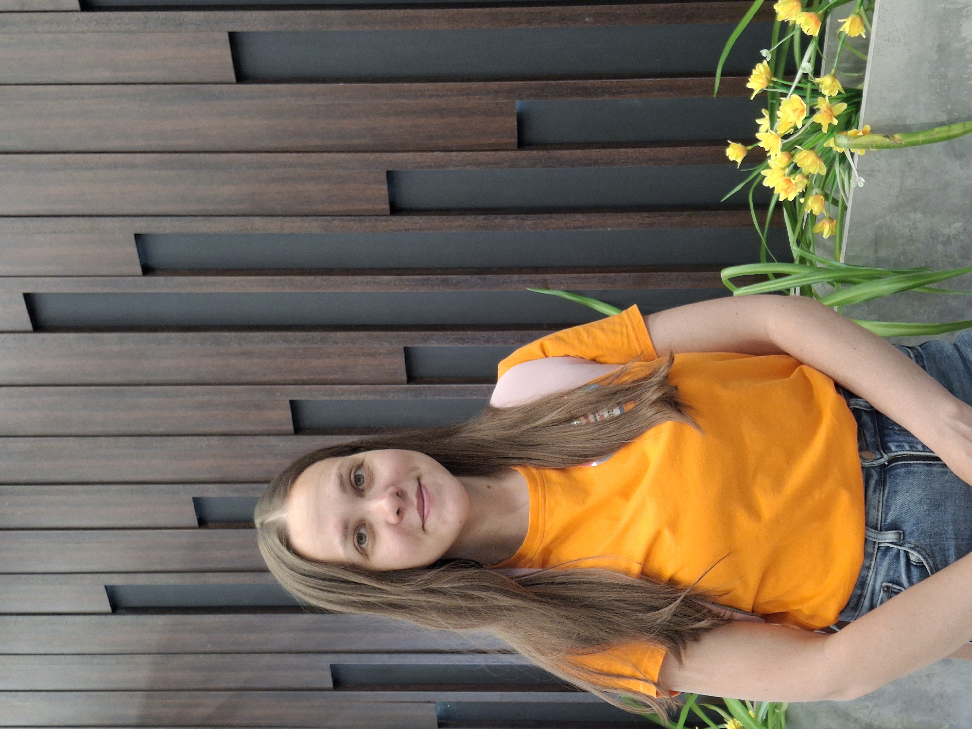 A woman with long, light brown hair is wearing an orange t-shirt and blue jeans. She's sitting in front of a decorative wooden wall with narrow, horizontal planks of dark brown wood separated by a recessed dark gray background. To her right, there's a vase with yellow flowers and green leaves.
The location is Chaikovsky, Russia.