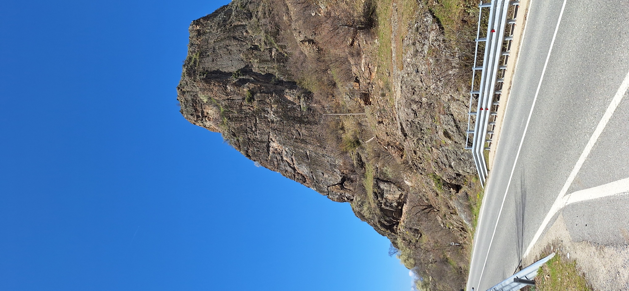 The image captures a roadside scene in Rila, Bulgaria. A large, rocky cliff face rises abruptly beside the asphalt road, protected by a silver guardrail. The sky above is a clear, unblemished blue. The cliff itself is textured and layered, with sparse vegetation clinging to its surface. A utility pole is also visible on the cliff face. The road markings indicate a two-lane road, with solid white lines separating the lanes.
