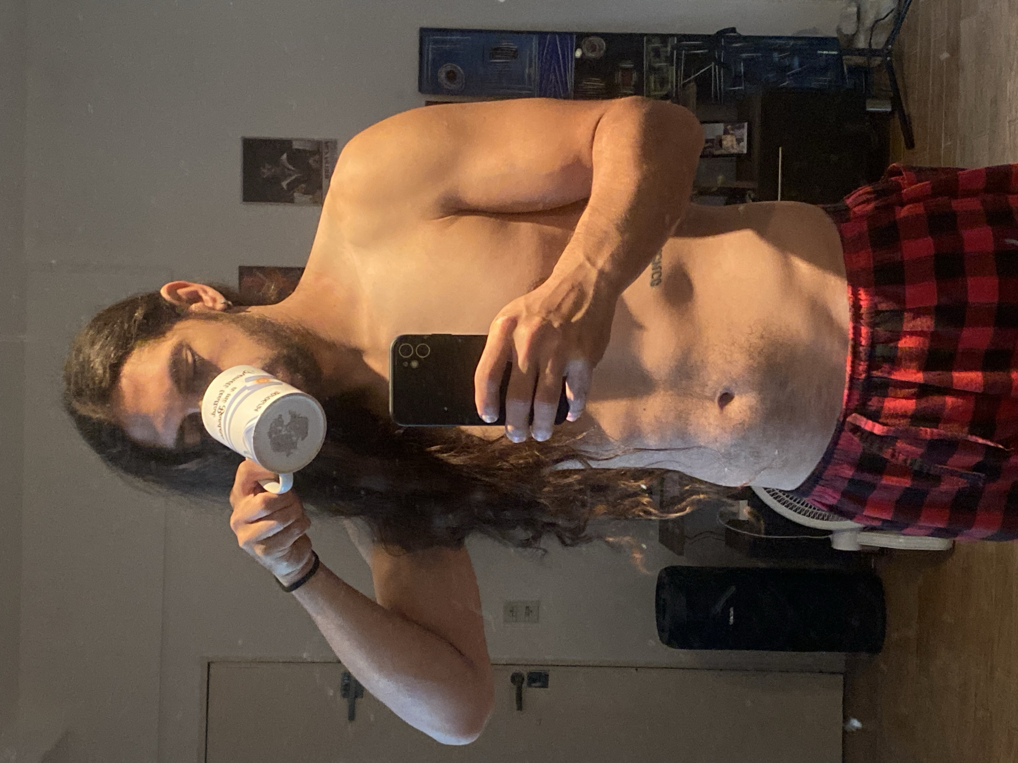 The image depicts a male individual captured in a mirror selfie within an indoor residential setting in Buenos Aires, Argentina. The subject possesses long, dark, wavy hair and a beard, and is shirtless, revealing a moderately muscular upper body. He is wearing red and black plaid pajama bottoms. A black bracelet adorns his left wrist, and a silver hoop earring is visible in his left ear. A mirrored inscription or tattoo is present on his left abdomen.
The individual is actively engaged in two simultaneous actions: holding a white mug with a mirrored design to his mouth with his left hand, appearing to drink, and holding a black smartphone with dual visible camera lenses in his right hand, pointing it towards the mirror to capture the reflection.
The background, visible through the mirror, features a light-colored wall. To the left, a light-colored door with a dark handle is partially reflected. On the floor in the mid-ground, a tall, black cylindrical object stands beside a white oscillating fan. Further to the right, a dark piece of furniture is visible, above which several wall-mounted items, including an abstract blue and black artwork, are reflected. The flooring throughout the visible area is wooden or laminate, and power cables are present on the floor in the far right portion of the frame.
