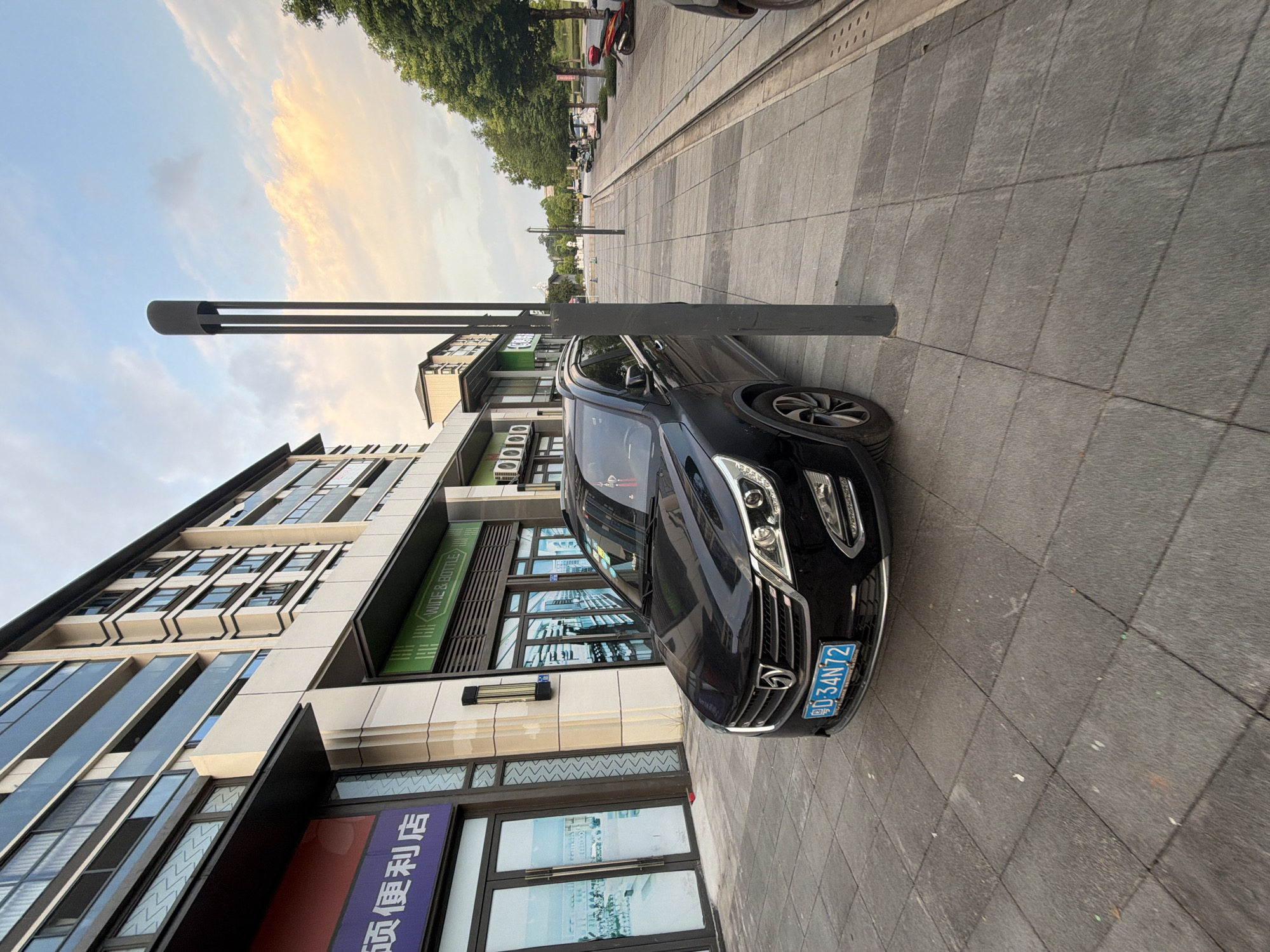 A black SUV is parked on a tiled sidewalk in front of a modern building with glass windows. The building appears to be a commercial establishment, with signage indicating "WINE & BOTTLE" and another sign with Chinese characters. A tall, dark lamppost stands near the car. The sidewalk is bordered by a street with trees lining the other side. The sky above is partly cloudy with hints of sunset colors. The license plate of the car reads "D-34N72". The scene is likely located in the city of Jiangjin, China, given the context provided.