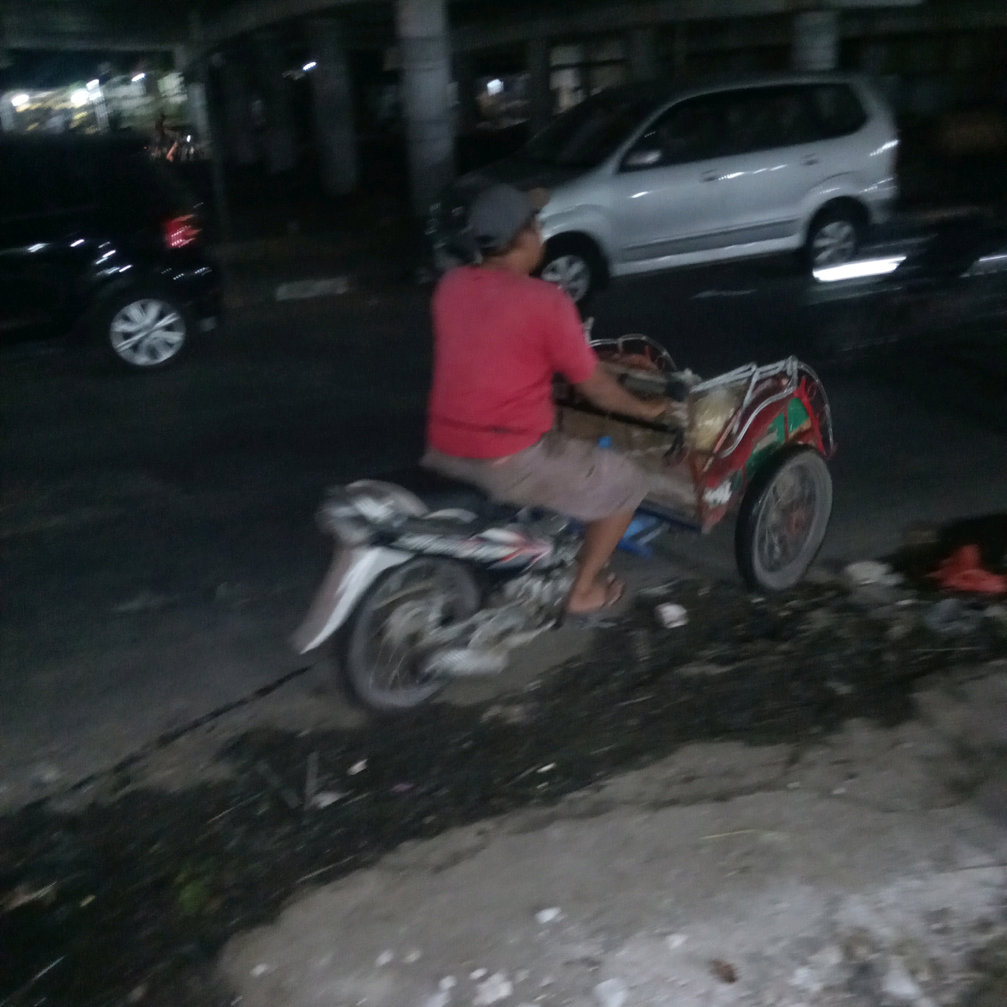 In Cileungsi, Indonesia, at night, a person wearing a red shirt and a gray cap rides a three-wheeled motorcycle. The motorcycle has a sidecar with red and green decorations. The scene is dimly lit, with a black car visible on the left and a silver van in the background. The ground is wet and appears to be a road or parking area, with some debris scattered around.
