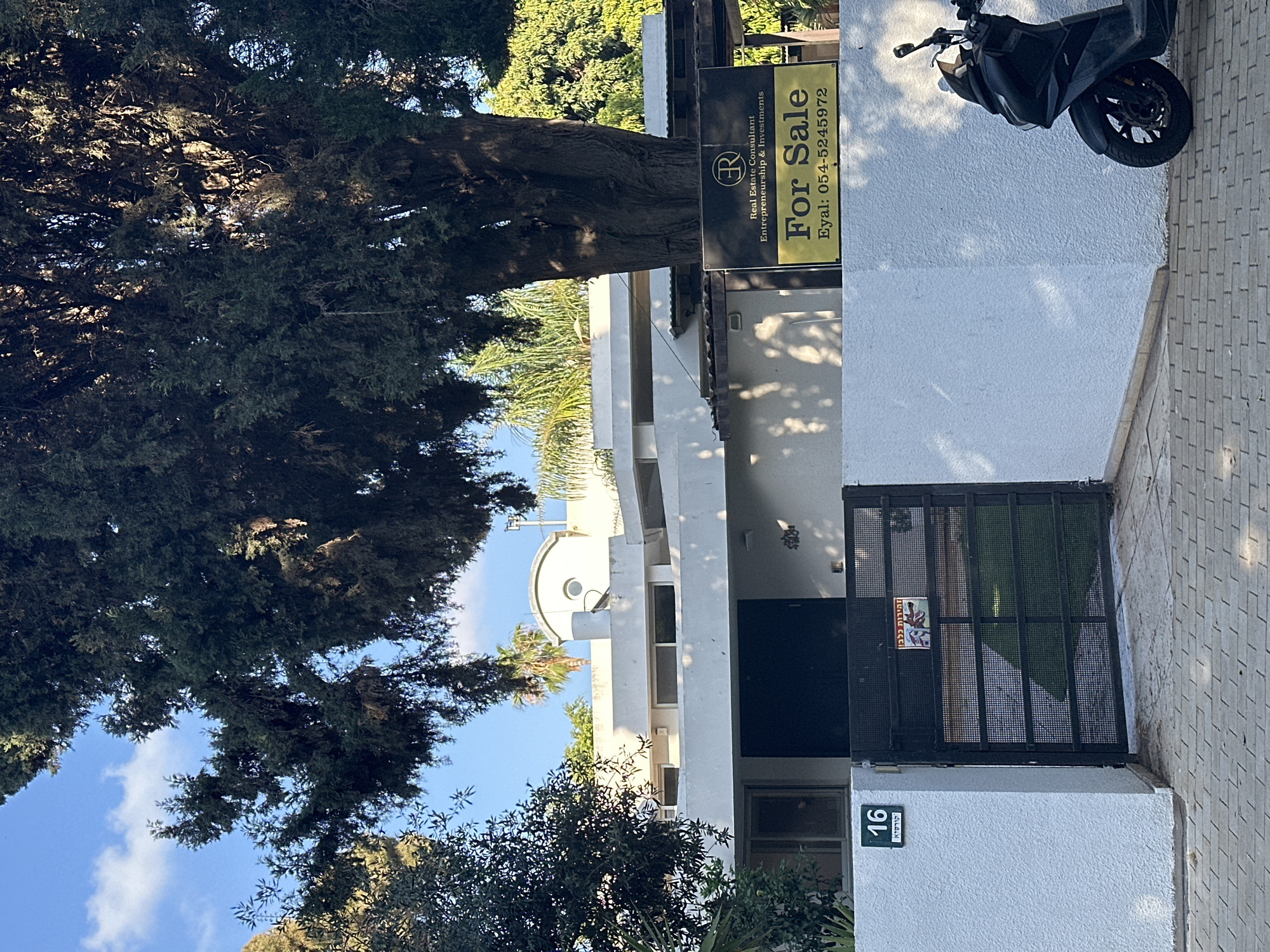 This image, taken during the daytime under a clear blue sky with scattered clouds, displays the exterior of a residential property. A prominent "For Sale" sign is visible, reading "R Real Estate Consultant Entrepreneurship & Investments For Sale Eyal: 054-5245972". The sign is affixed to a pole near a large, ancient-looking tree with dense green foliage that dominates the left side of the frame.
The house itself is predominantly white, featuring dark-framed windows and a sturdy black metal gate at the entrance. On the white wall beside the gate, a number plate clearly shows "16 קורסיא" (16 Kursia), identifying the house number and street name. A smaller, red and white sign on the gate warns "זהירות כלב!" (Caution Dog! in Hebrew).
A black scooter is parked on the light-colored, patterned paved area in front of the house, against a white wall. The scene is well-lit, with distinct shadows cast on the house and ground, indicating direct sunlight. Various green trees and bushes surround the property, adding to a lush environment, including what appears to be a palm tree in the background. The visible Hebrew text and phone number with an Israeli prefix suggest the location is in Israel, specifically the street Kursia.