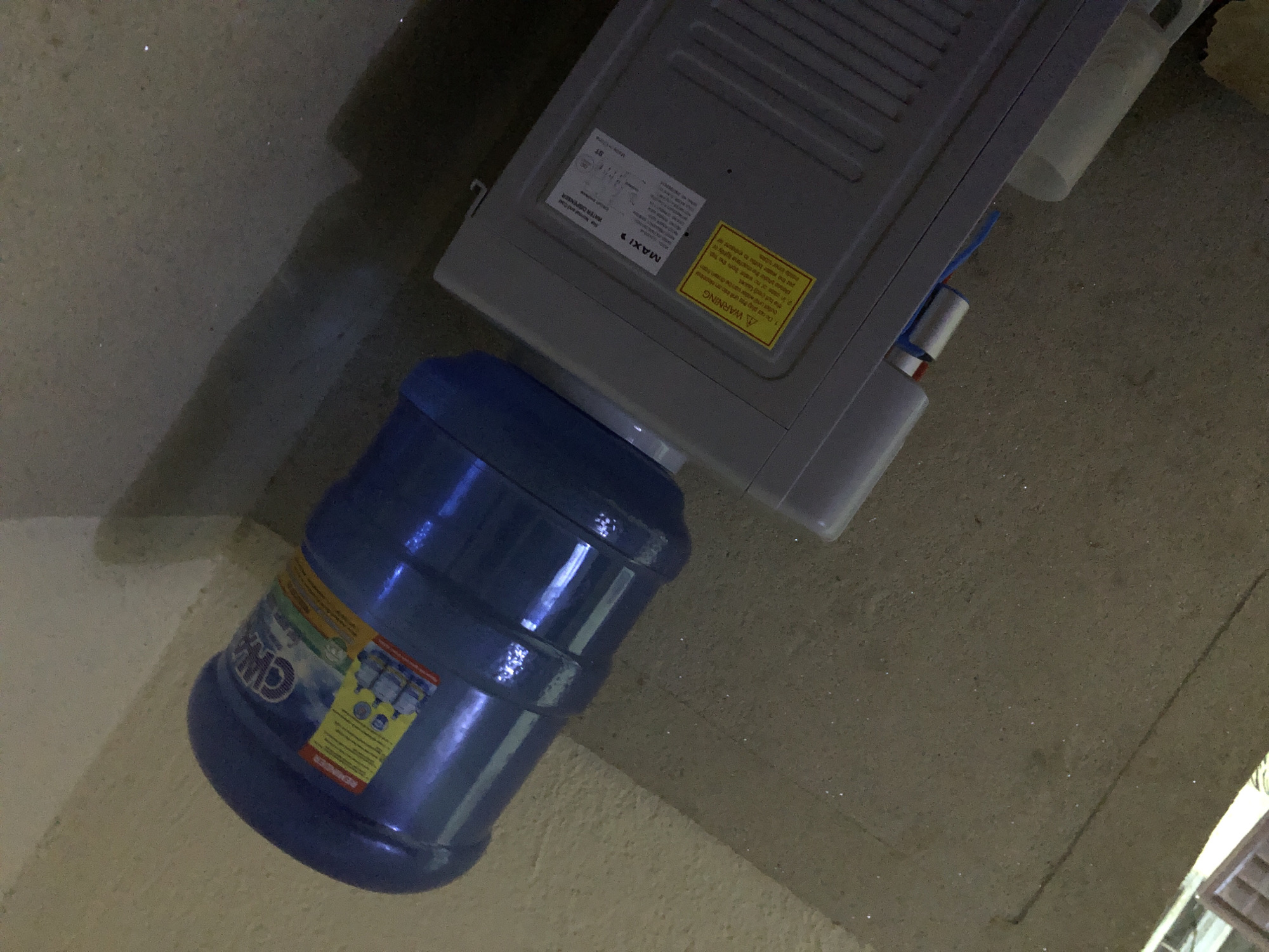 In Kabong, Nigeria, a blue water cooler bottle is placed on top of a gray water dispenser. The water bottle is larger than the dispenser, with its base resting on the top surface of the dispenser. The label on the water bottle is partially visible, showing text and images. The water dispenser has a metallic grille on its side and a label with text. A yellow warning sticker with text in black is also visible on the dispenser. Wires and a pipe are connected to the dispenser. The scene is set against a textured, concrete-like wall and floor, with shadows cast by an unseen light source.