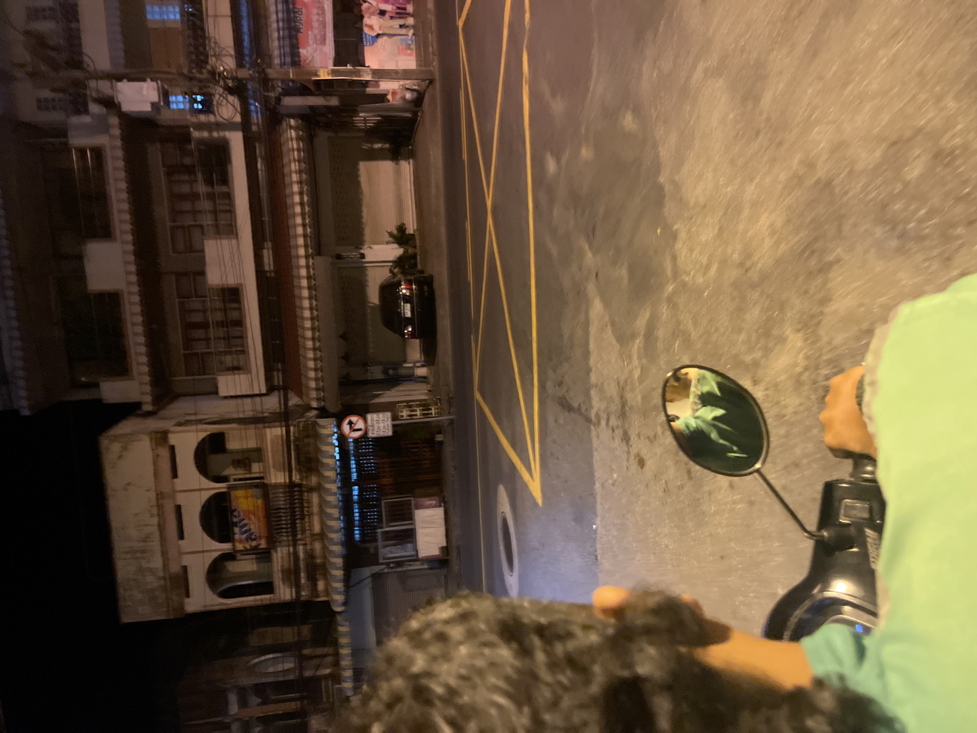 A POV shot from a motorcycle at night in Phuket City Municipality, Thailand, shows a street scene. The foreground is dominated by the back of a person's head with dark, curly hair and a bright green shirt, holding the handlebars of a scooter. The mirror reflects some foliage and possibly a person. The street has yellow lane markings and a circular manhole cover. In the background, buildings line the street, with a black car parked in front of one. A sign with a red arrow and Thai text is visible. The lighting suggests a commercial or residential street.