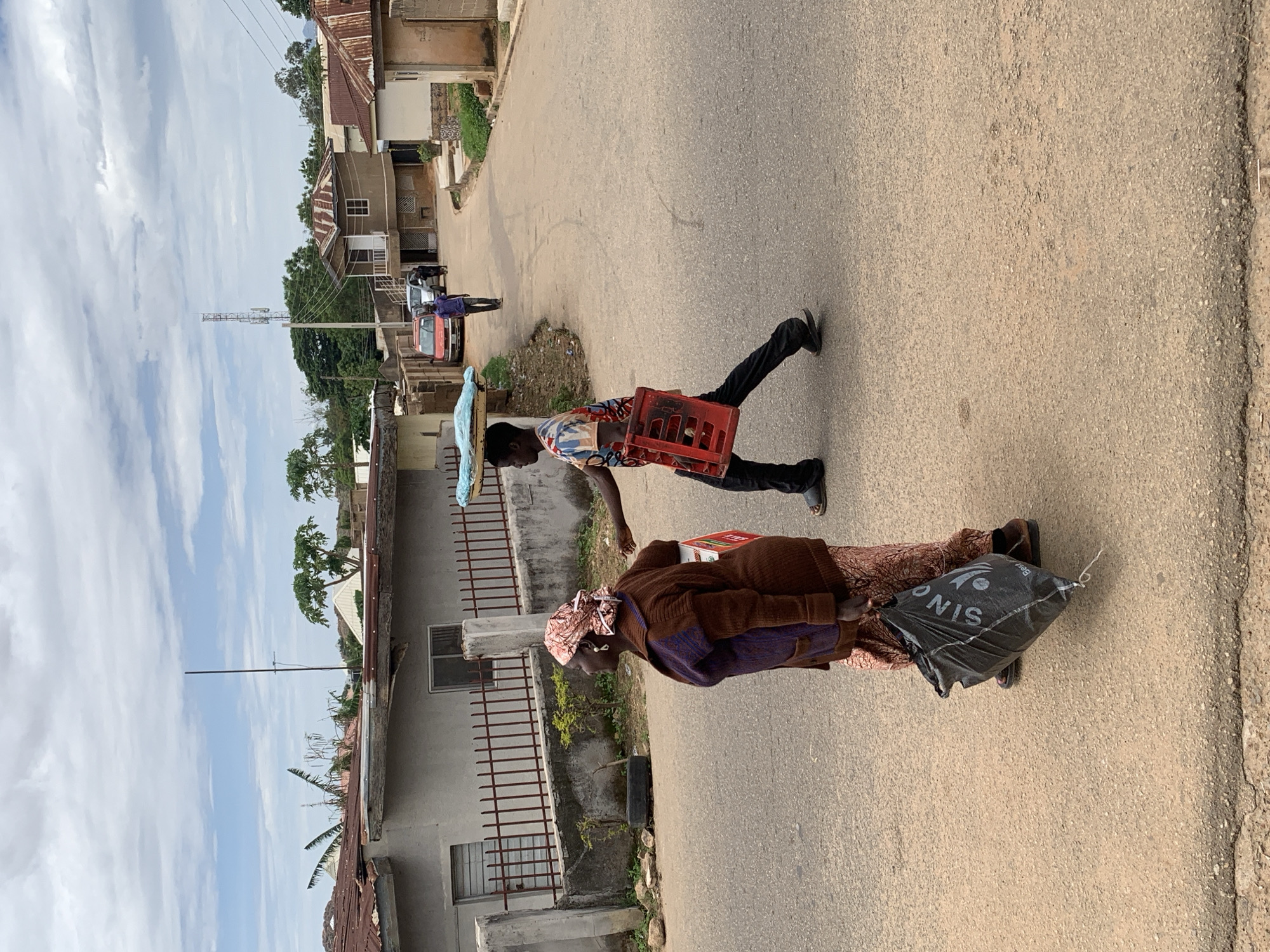 The image depicts a street scene in Jos, Nigeria, under a predominantly cloudy sky. The foreground and mid-ground are dominated by an asphalt road that appears worn.
In the left foreground, an older woman with dark skin walks towards the left, looking slightly left. She wears a brown long-sleeved cardigan with purple accents, a light-colored patterned skirt, and a patterned headwrap in similar colors. She carries a black plastic bag in her left hand, which has partial white lettering, "SINO," visible.
Slightly to the right and further back, a younger man with dark skin walks towards the right. He wears a multi-colored short-sleeved t-shirt, dark trousers, and dark flip-flops. He carries an empty red plastic crate in his left hand, held against his side, and balances a light blue, possibly fabric-wrapped, object on his head.
In the background on the left, a light-grey concrete building with a rusty corrugated metal roof is partially visible behind a dark red metal fence. The building features two rectangular windows, one with horizontal bars. A discarded black tire rests on the ground near the fence. Above this building, a thin, dark vertical pole, possibly an antenna, extends upwards.
The road curves to the right in the mid-distance, where several houses with light-colored walls and rusty corrugated metal roofs line both sides. A distinct two-story house with a rounded base and a small balcony is visible on the right. Two vehicles are parked on the right side of the road: a red sedan followed by a silver car. Further down the curving road, a third individual, dressed in a purple shirt and dark trousers, walks away from the viewer.
A tall metal telecommunications tower is visible in the far background on the right, above the houses and trees. Scattered green trees and sparse vegetation are present throughout the background.