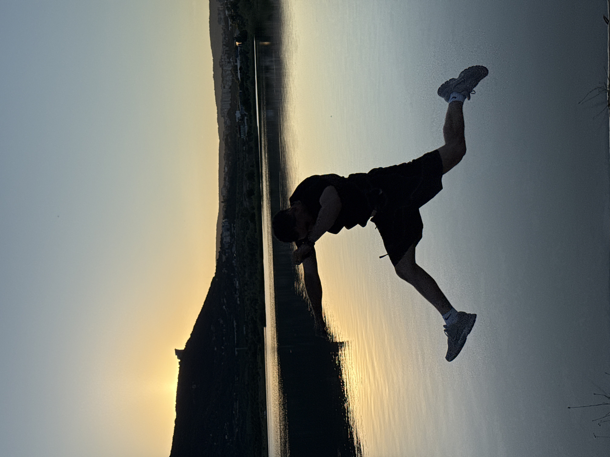 A silhouetted man is captured mid-air, jumping over a body of water at sunset. The man is wearing a t-shirt and shorts, with his arms bent and body angled as if in a dynamic pose. His sneakers are visible as he extends his legs. The water reflects the warm hues of the sky, with the sun low on the horizon, casting a golden glow. In the background, a dark, tree-covered hill is silhouetted against the sky, with buildings visible on the distant hills, suggesting an urban or suburban environment. This scene appears to be taking place at the artificial lake located on the western outskirts of Shumen, Bulgaria.