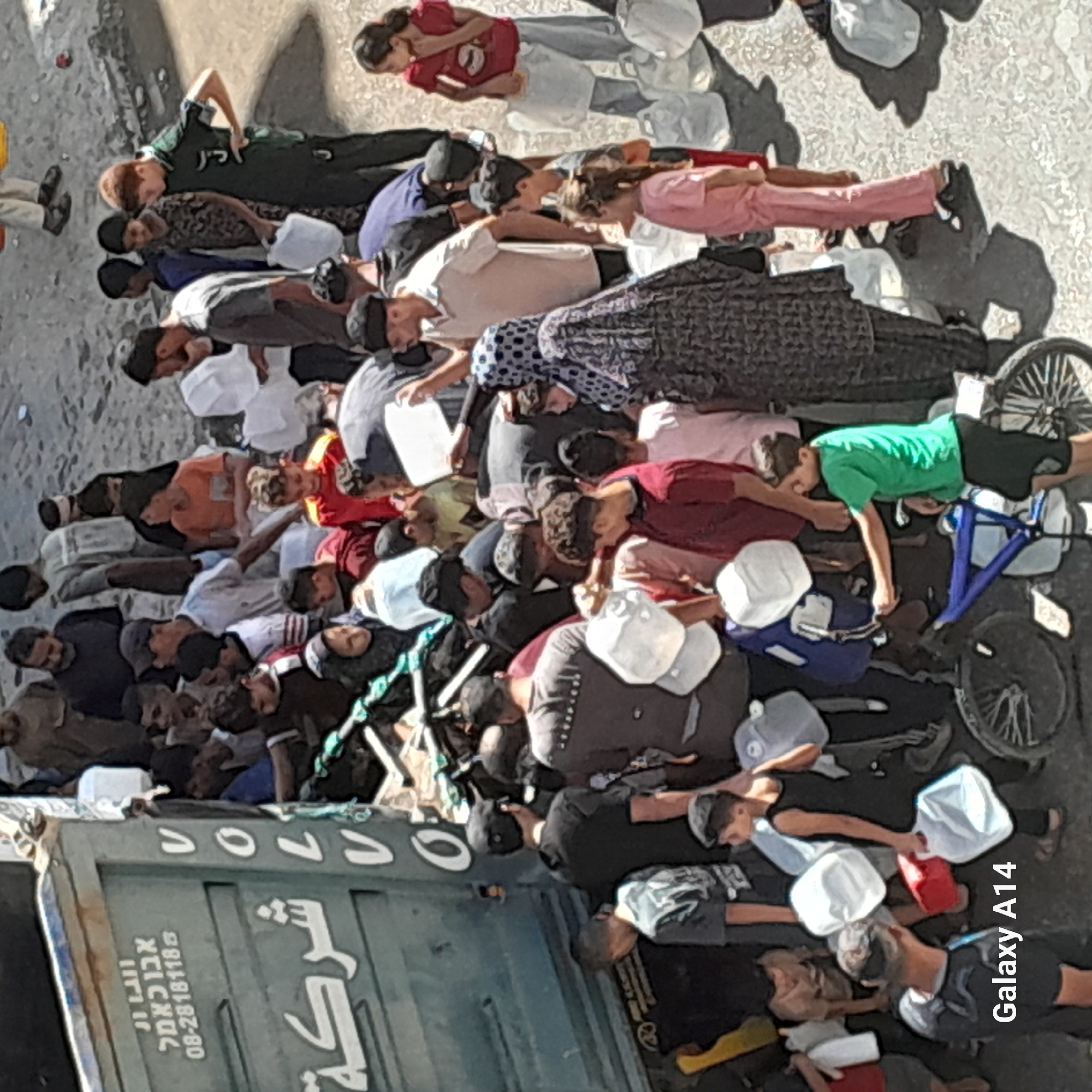 In an outdoor urban setting, a group of people, primarily men and boys, are gathered around the open back of a large, dark green truck, possibly a delivery vehicle. Some individuals are reaching into the truck, while others appear to be receiving or holding large, white plastic containers, some resembling jerrycans or water jugs. A few red and blue plastic containers are also visible. The ground is paved with rough, light-colored material, and shadows indicate a bright, possibly sunny day. The truck has some text and logos on its side, including what appears to be Hebrew lettering and the Volvo logo. The people are dressed in casual clothing, with some wearing traditional head coverings. The overall scene suggests the distribution or collection of essential supplies, such as water, in a public space.