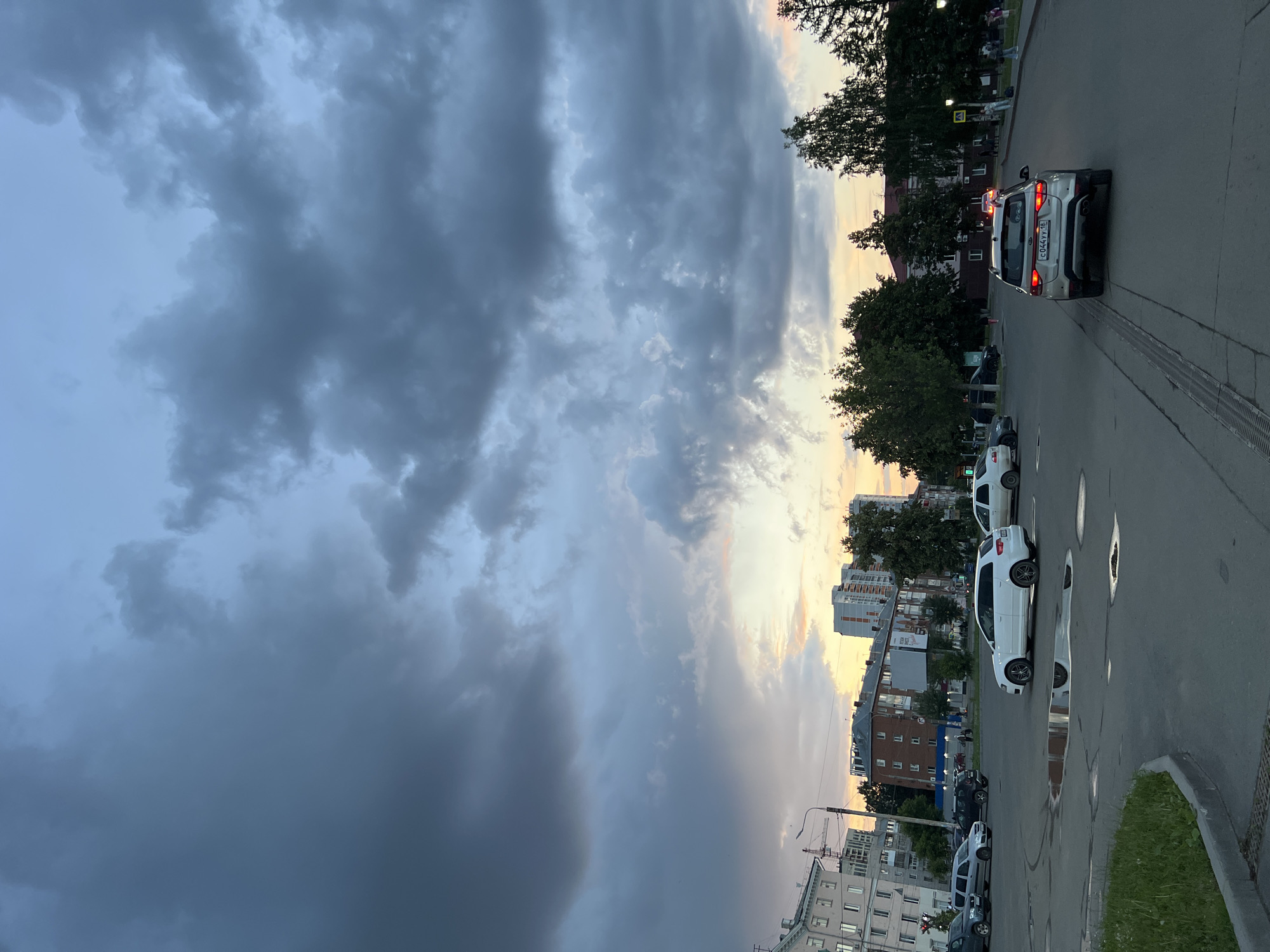 The image shows a street scene in Izhevsk, Russia, with a dramatic cloudy sky overhead. In the foreground, a light-colored SUV is driving away from the camera on a paved road. The SUV has a license plate that reads "C 044YY 18". To the left, a white hatchback is parked on the street. Several other cars are parked or driving in the background. The street appears wet, with several puddles visible on the asphalt. Buildings, including apartment complexes and trees, line the street. The sky is filled with dark, heavy clouds, with hints of a colorful sunset peeking through in the distance. The overall impression is of a city preparing for or recovering from rain, with an ominous and beautiful sky.