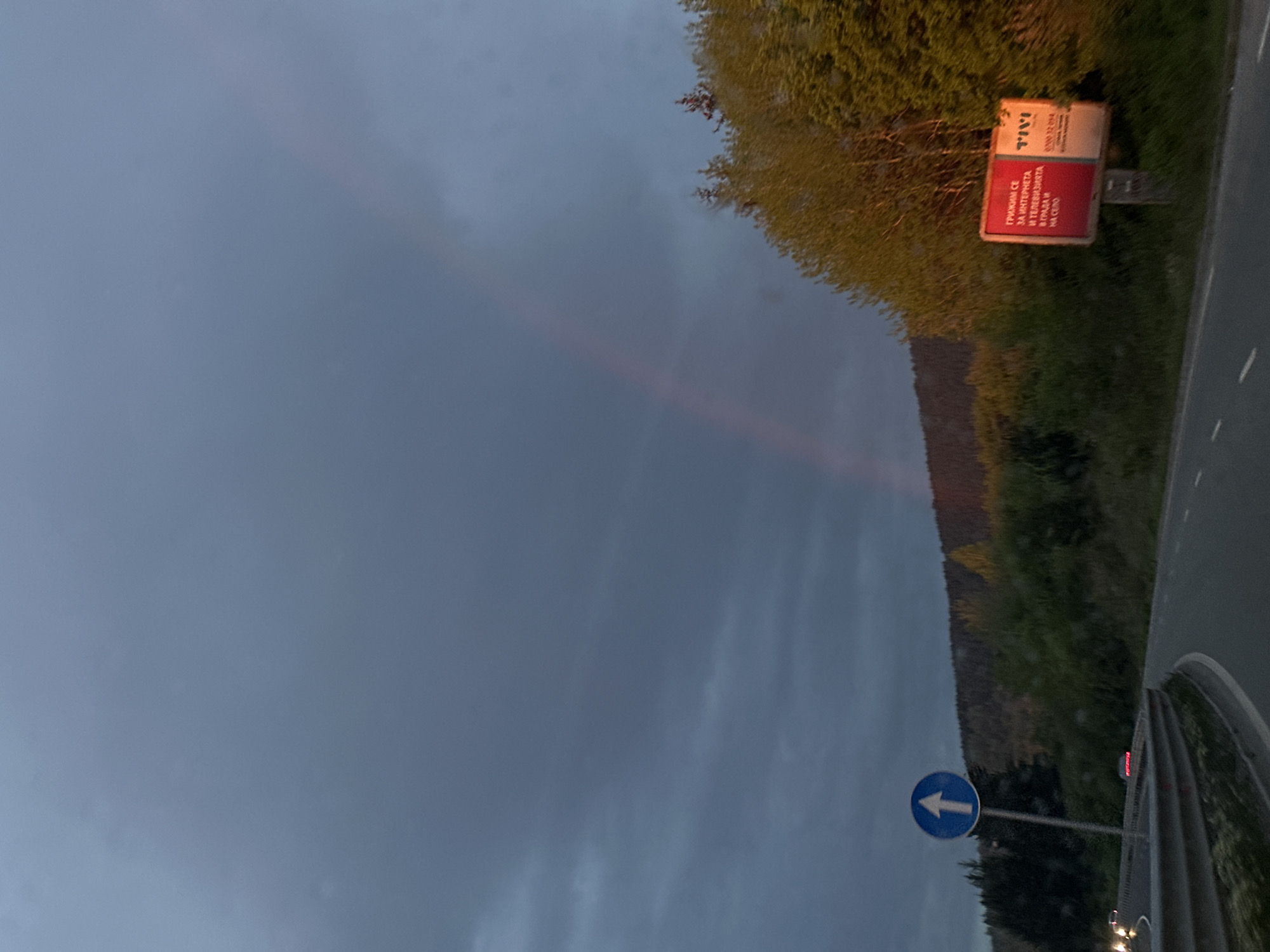 The image presents a view of the sky and roadside in what appears to be a rural area. A faint rainbow extends across the sky. The presence of roadside vegetation, a road, and signs, one of which reads "TVIVI" and another a blue directional sign, suggest the image was taken along a road. The text on the red sign appears to be in Bulgarian, suggesting a location in Bulgaria. It is unclear that the location is in Marchaevo. Further specifics would require more detailed analysis and potential investigation.