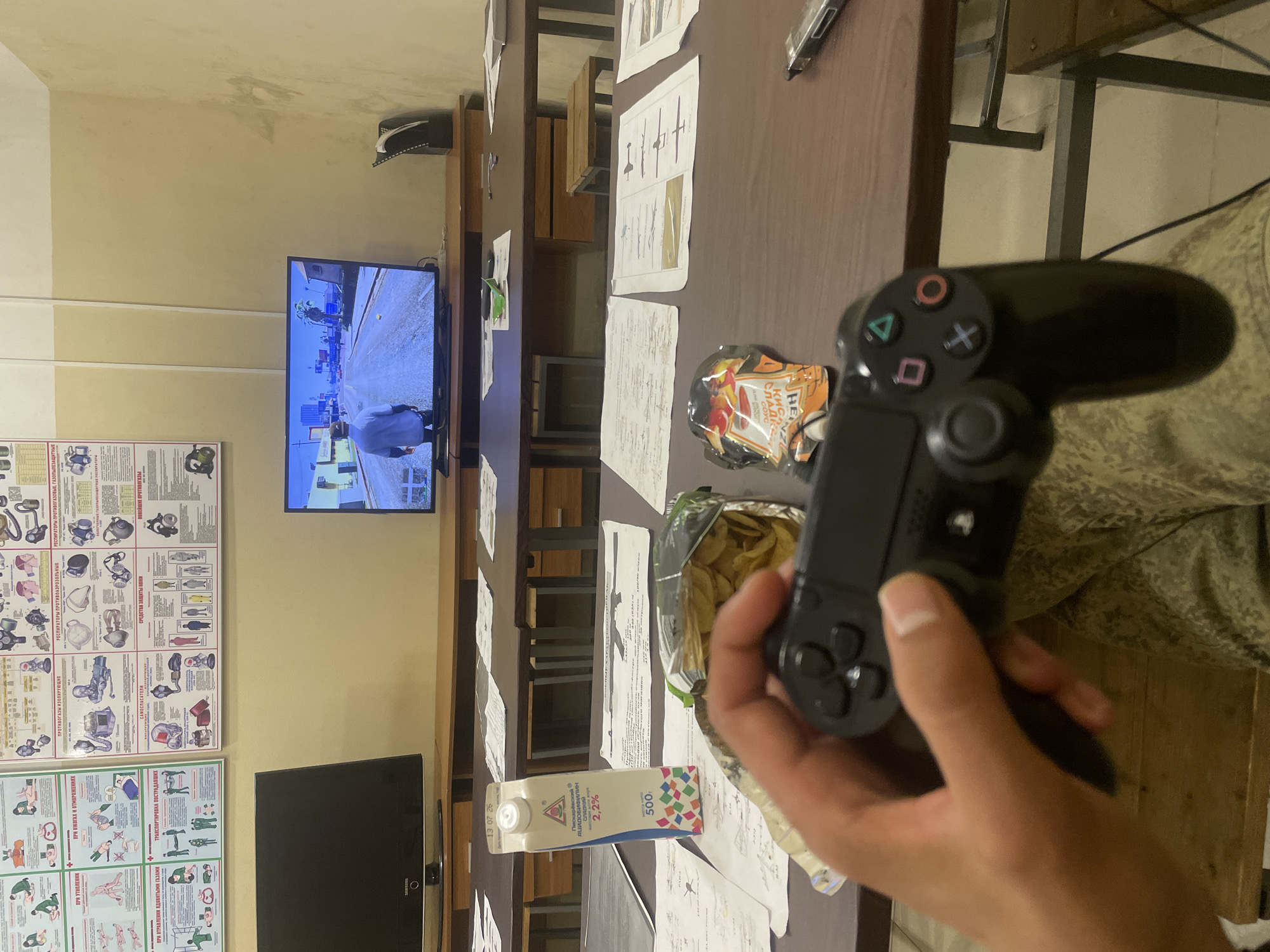 The image depicts a scene indoors, likely in a classroom or communal area, with a person holding a video game controller and playing a game on a television screen. The television displays a scene from the video game Grand Theft Auto V, showing a character walking down a street in a city. On the table in front of the person are a carton of milk and bags of snacks, suggesting a casual leisure activity. The room appears to be furnished with desks and chairs, and educational posters are visible on the wall, some of which detail safety procedures or technical information. The overall atmosphere is relaxed and informal. The location is identified as Оредеж, Russia.
