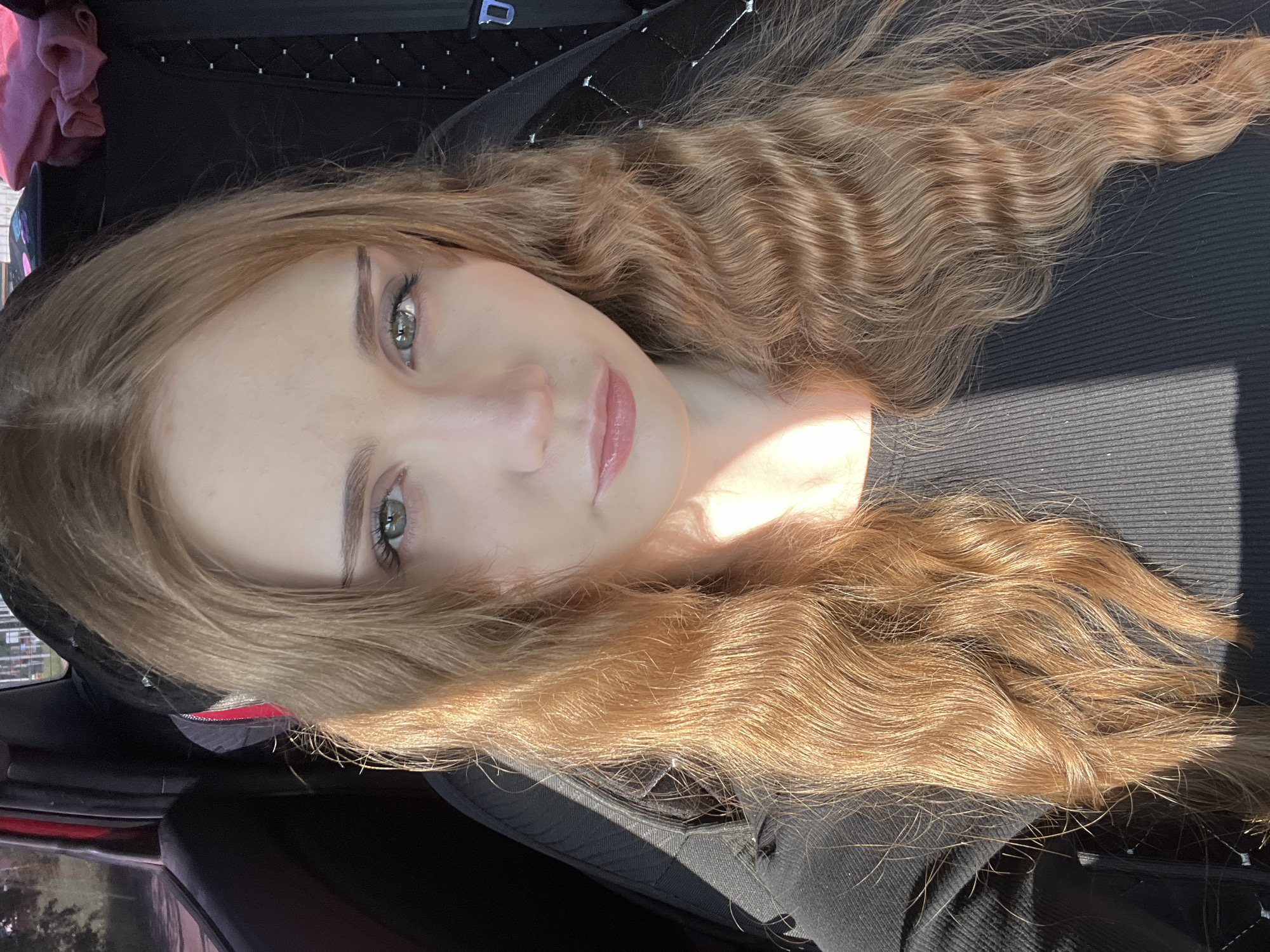 A young Caucasian woman with long, wavy, light brown hair is shown in a close-up, selfie-style photograph taken from inside a vehicle. Her eyes are light green and she is wearing a black, ribbed, long-sleeved top. Sunlight casts a shadow diagonally across her face and upper body. The background shows the interior of a car, with a black seat and a red interior door panel visible. There is no indication of the specific location within Загородный, Russia, other than the general context of being inside a vehicle.