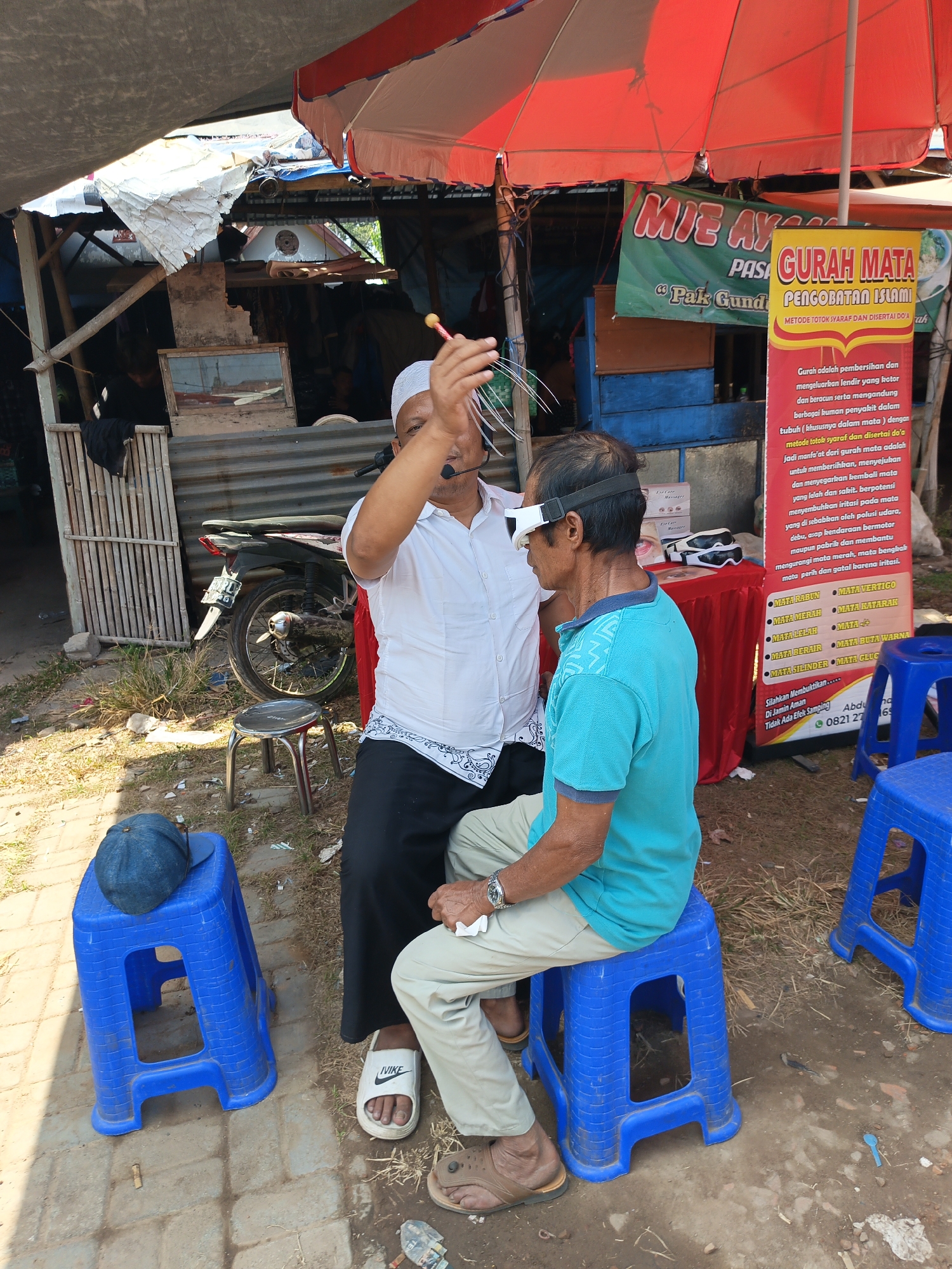 In Jatinom, Indonesia, an outdoor scene unfolds under a red and white umbrella. A man, wearing a white shirt and a white cap, is in the process of performing an eye treatment on another man. The man receiving treatment wears a teal polo shirt and a white device around his eyes. He is seated on a blue plastic stool, while the practitioner stands. In the background, a motorcycle is parked near a corrugated metal structure. A banner, displaying text in Indonesian, is visible behind the seated man, advertising "GURAH MATA" and "PENGOBATAN ISLAMI" (Islamic Eye Treatment). Several other blue plastic stools are scattered around the area. The ground is a mix of dirt and paved bricks, with dry grass and debris present.