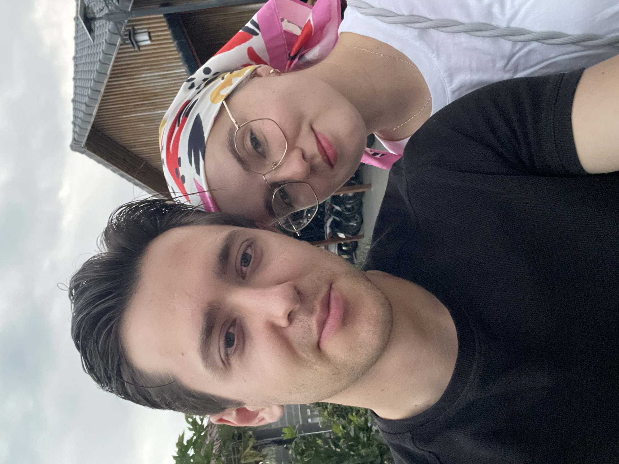 A photograph shows two people, a man and a woman, in an outdoor setting. The woman is wearing a white, patterned headscarf and round-framed glasses. The man is wearing a black shirt. Both are positioned close to each other. The background includes a building with a roof and glimpses of greenery. The location is Phú Quốc, Vietnam.