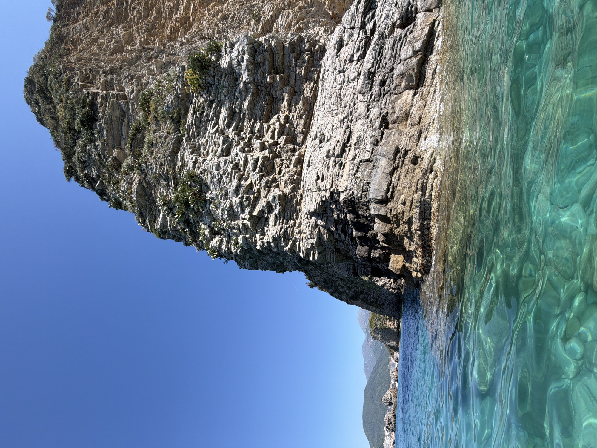 The image depicts a rocky cliff face meeting the turquoise water of the Adriatic Sea. The cliff is composed of jagged, gray stone formations with sparse vegetation clinging to its surface. The sea water is clear enough to see the rocks beneath the surface. In the distance, there are more cliffs and mountains. The sky is clear and blue. The image was taken in Budva, Montenegro, a city known for its coastline.