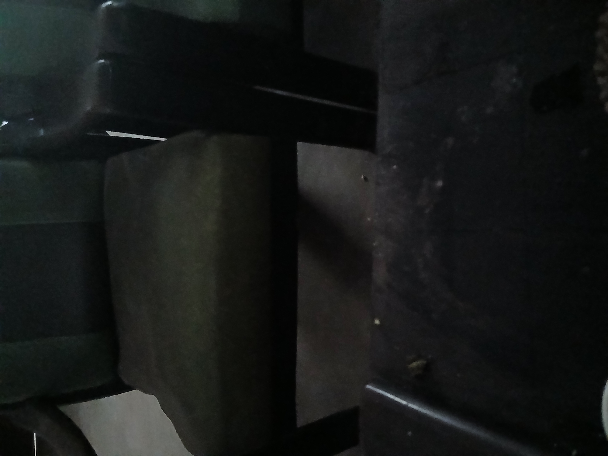 A dark, possibly worn, armchair is seen from a slightly elevated perspective. The armchair is upholstered in a dark fabric with a subtle striped pattern on the backrest. The seat is a darker shade of the same fabric. The right armrest is particularly prominent, appearing to be made of dark wood. The armchair is situated in an indoor environment, with a dark floor visible beneath it. In the foreground, a dark, flat surface, possibly a table, is visible, also appearing somewhat worn and dusty. The lighting in the scene is dim. The location is the city of Tenggarang, Indonesia.