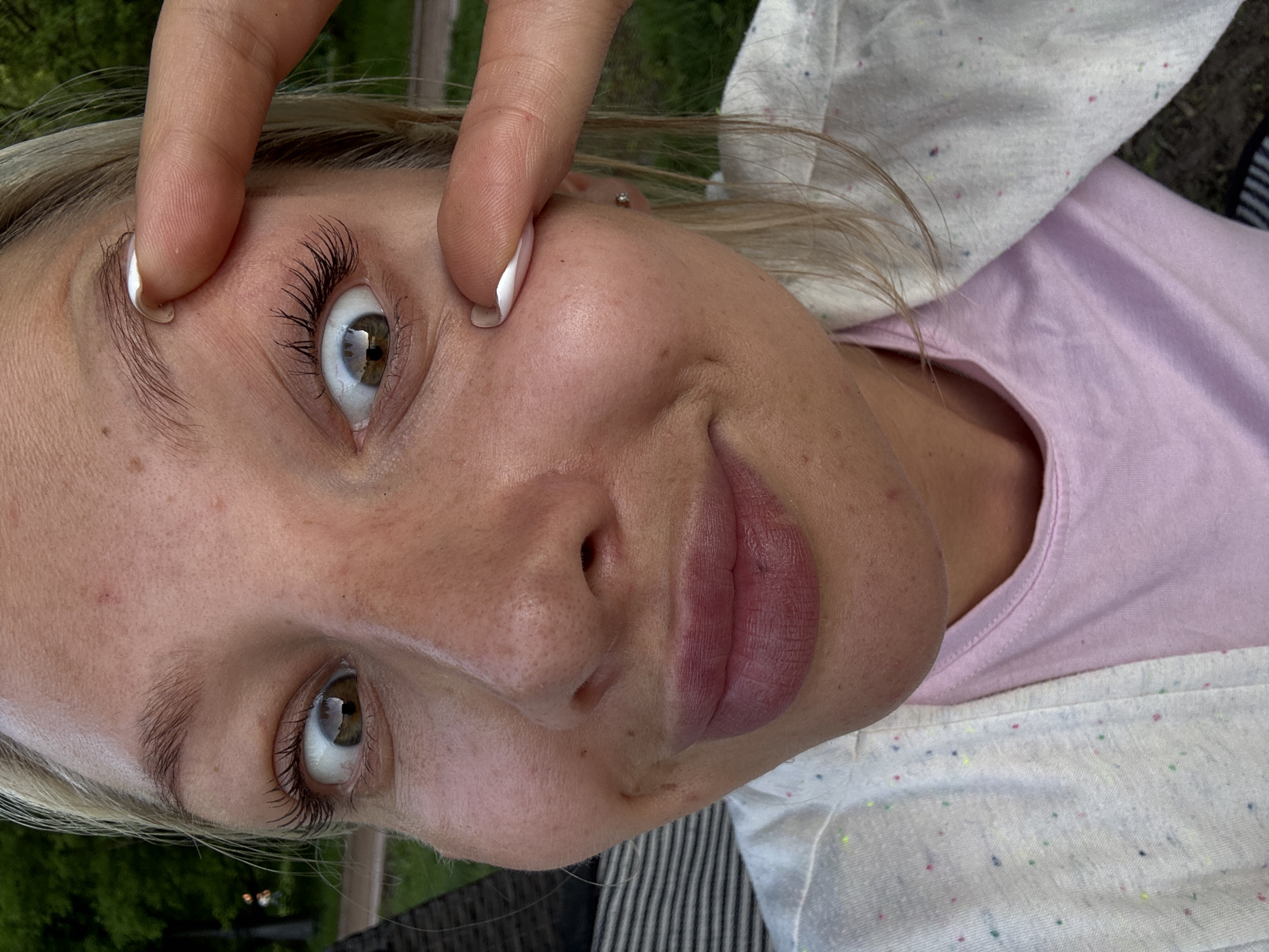 A close-up, tilted portrait photograph depicts a light-skinned woman with blonde hair. She is wearing a light pink t-shirt with a white cardigan over it. Her left eye is slightly widened and her other eye is held open by fingers. Her skin has a number of freckles and moles. The background shows greenery, hinting at an outdoor location. There is no visual evidence suggesting the location to be Moscow, Russia.